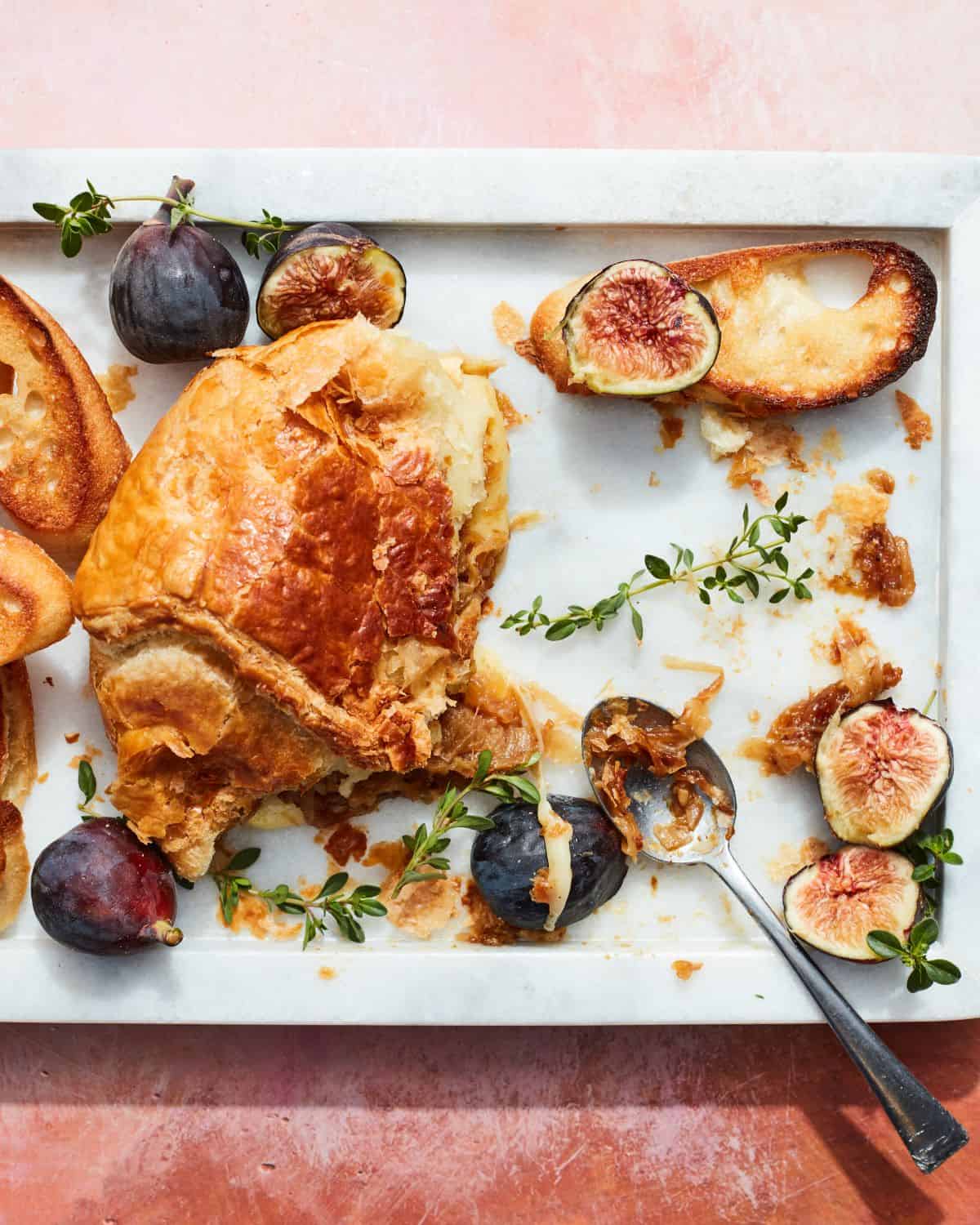 A white rectangular platter with figs, baguette slices, and some puff pastry wrapped brie leftovers along with thyme and caramelized onions and a spoon.