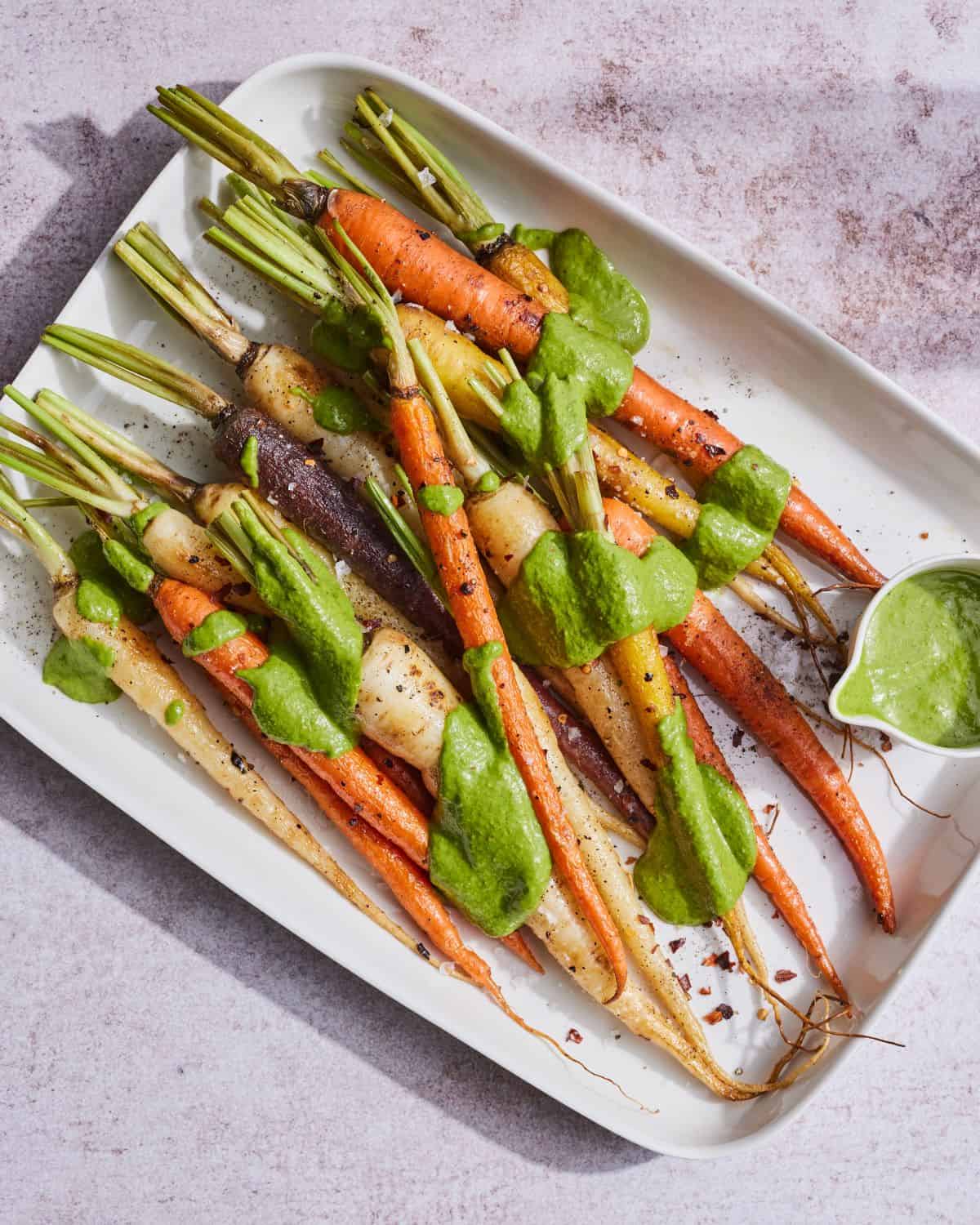A rectangular white platter with charred rainbow carrots with a drizzle of basil mint sauce along with a small bowl of the same sauce in the platter. 