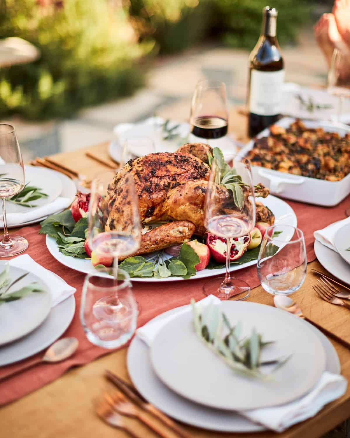 Dinner table with a whole roasted turkey plate in the center, wine glasses and plates, a baking dish with stuffing in the back and a bottle of wine.