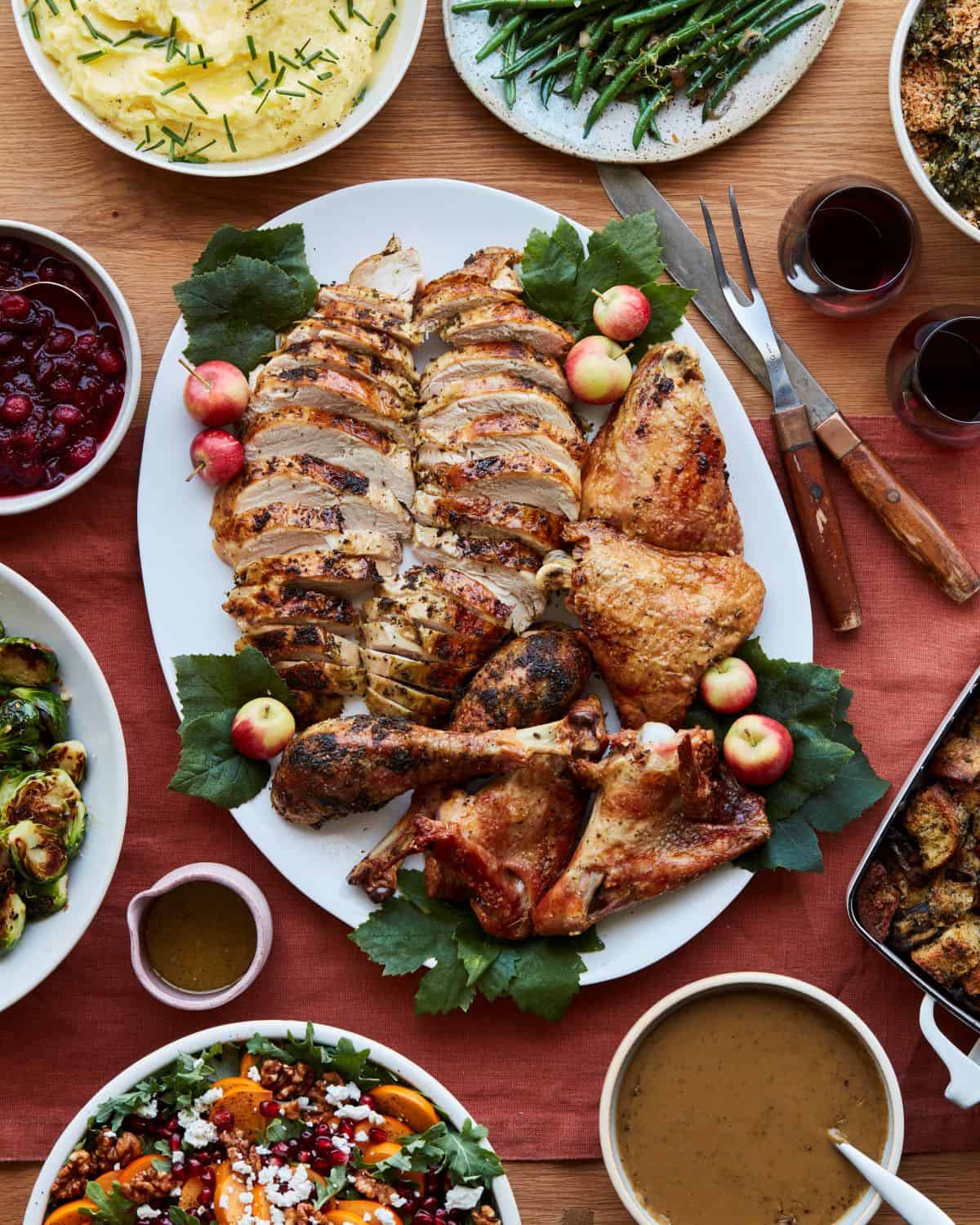A shot of a Thanksgiving dinner table from top, with a carved herb roasted turkey in a white plate in the center, with gravy, red wine, cranberry sauce, brussels sprouts, mashed potatoes, green beans and stuffing around it, and a carving fork and knife next to the turkey plate.