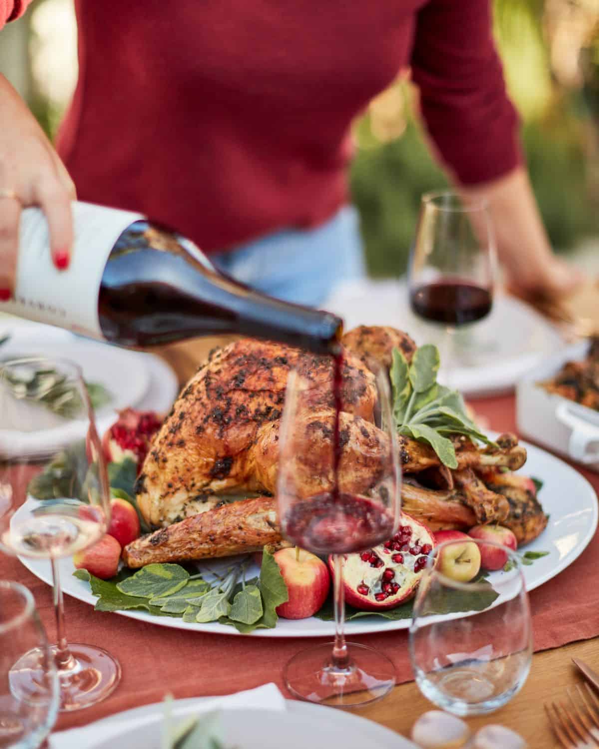 A dinner table with wine glases, plates, and a white plate with whole roasted turkey in it, with a blurry background of a woman pouring red wine into a wine glass.