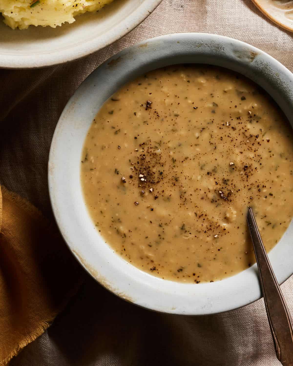 A close-up shot of a grey bowl with turkey gravy garnished with freshly ground black pepper with a spoon in it, with a bowl of mashed potatoes on the side.