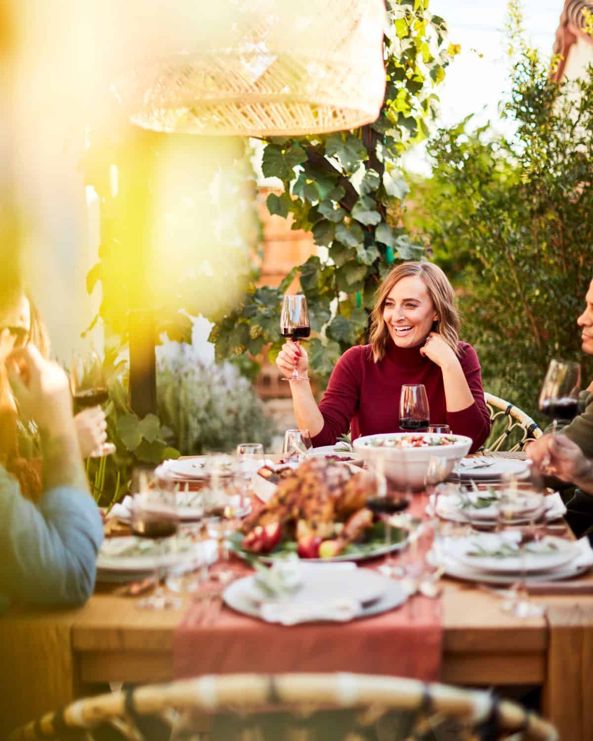 A dinner table for Thanksgiving with plates, wine glasses, a whole roasted turkey platter and a big bowl of salad in a blurry foreground with a clear background of a woman (Gaby) in a maroon sweater seated at the head of the table, raising a wine glass.