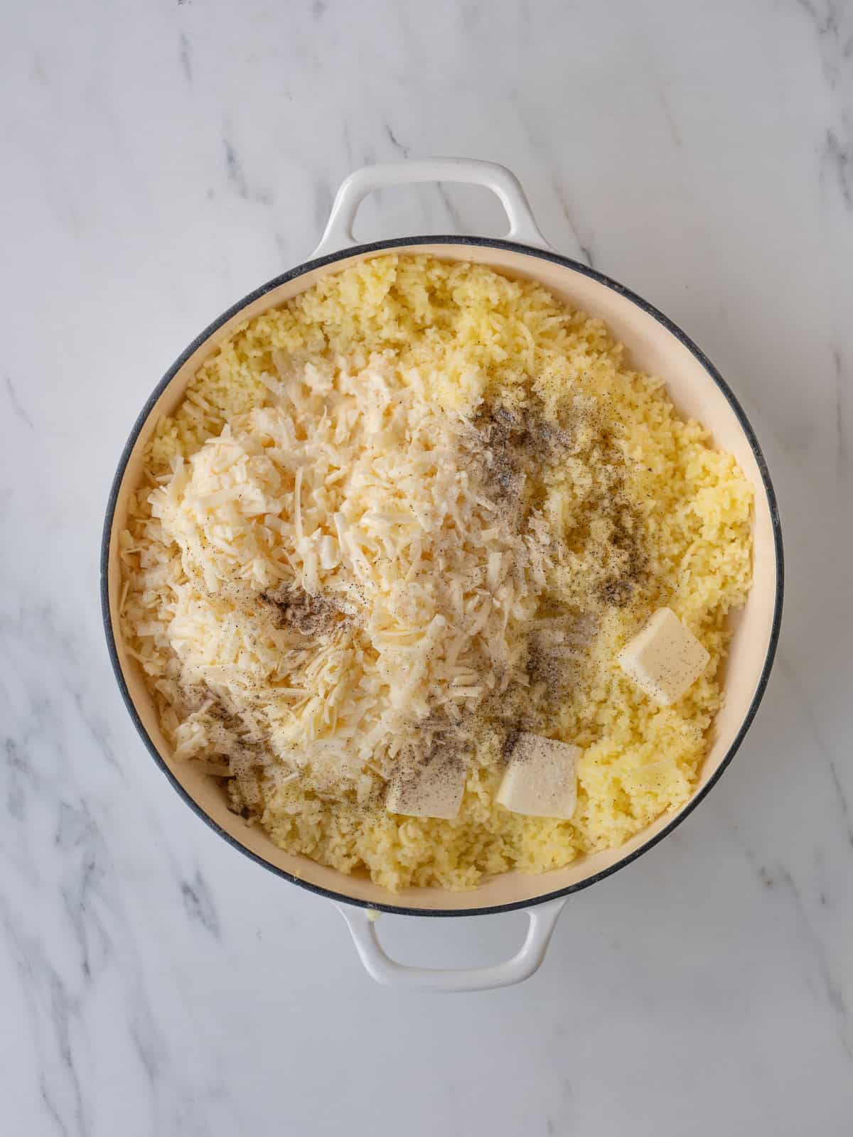 A dutch oven with shredded boiled potatoes, and cream, white cheddar, cubes of butter, salt and pepper added on top.