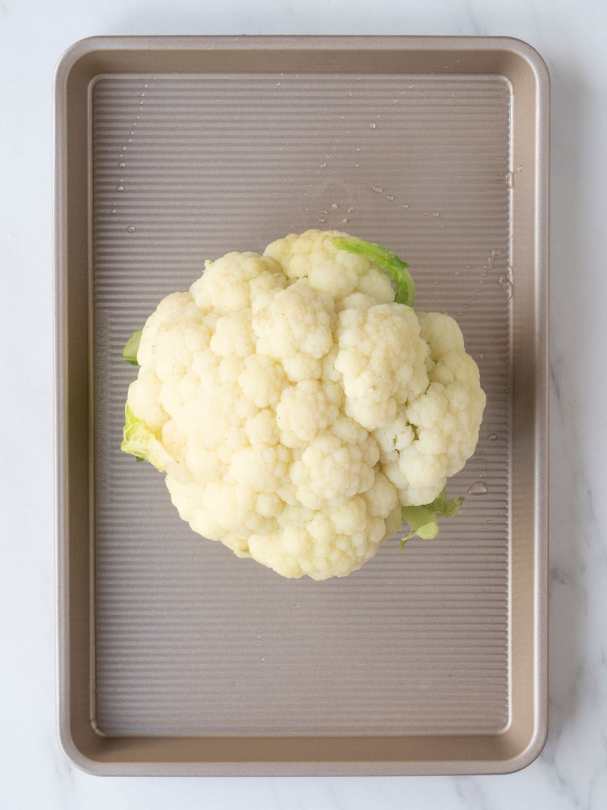 A sheet pan with a whole roasted cauliflower placed at the center, drizzled with olive oil and seasoned with salt.