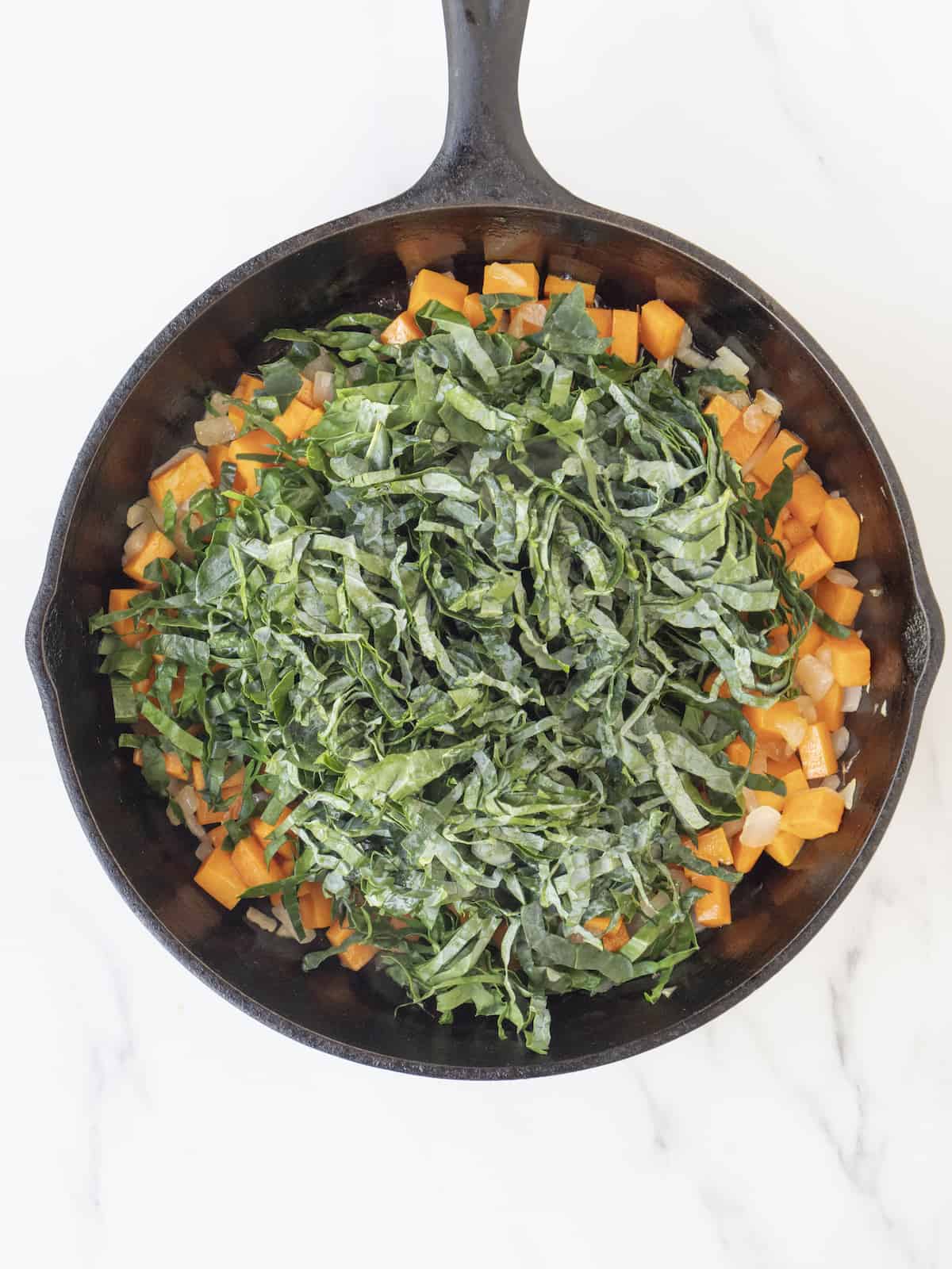 A skillet with cooked onions and sweet potatoes, topped with shredded kale and cooked down.