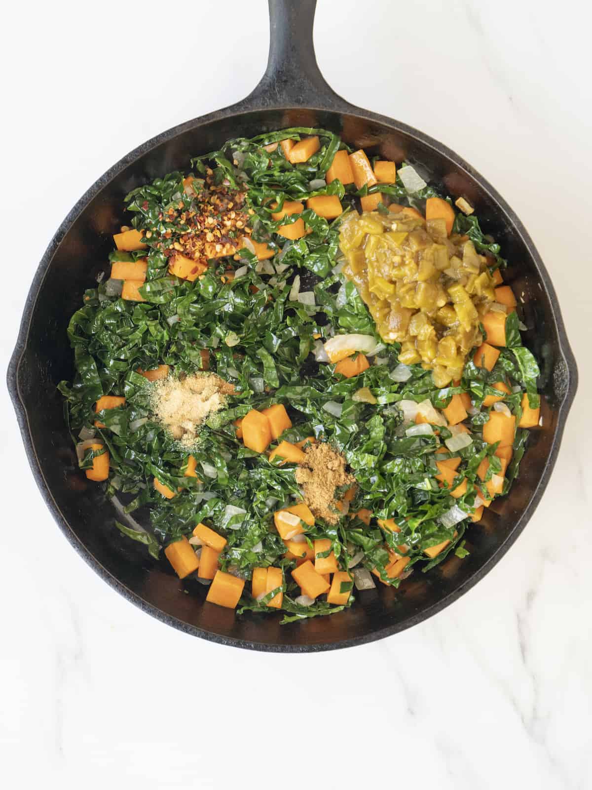 A skillet with cooked vegetables (kale, onions, sweet potatoes) topped with spices and diced green chiles.