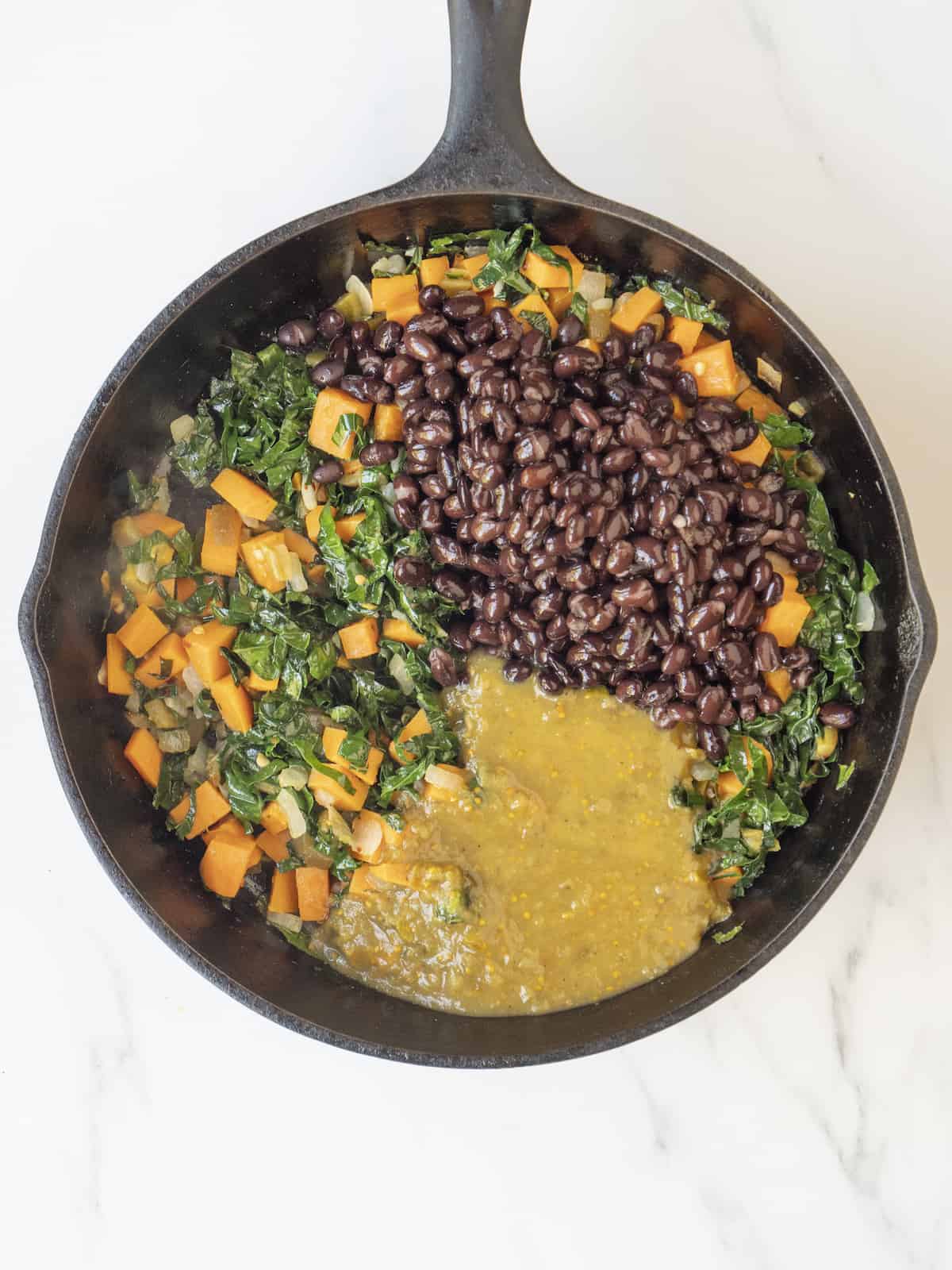 A skillet with cooked vegetables (kale, onions, sweet potatoes) topped with black beans and enchilada sauce, all being cooked together.