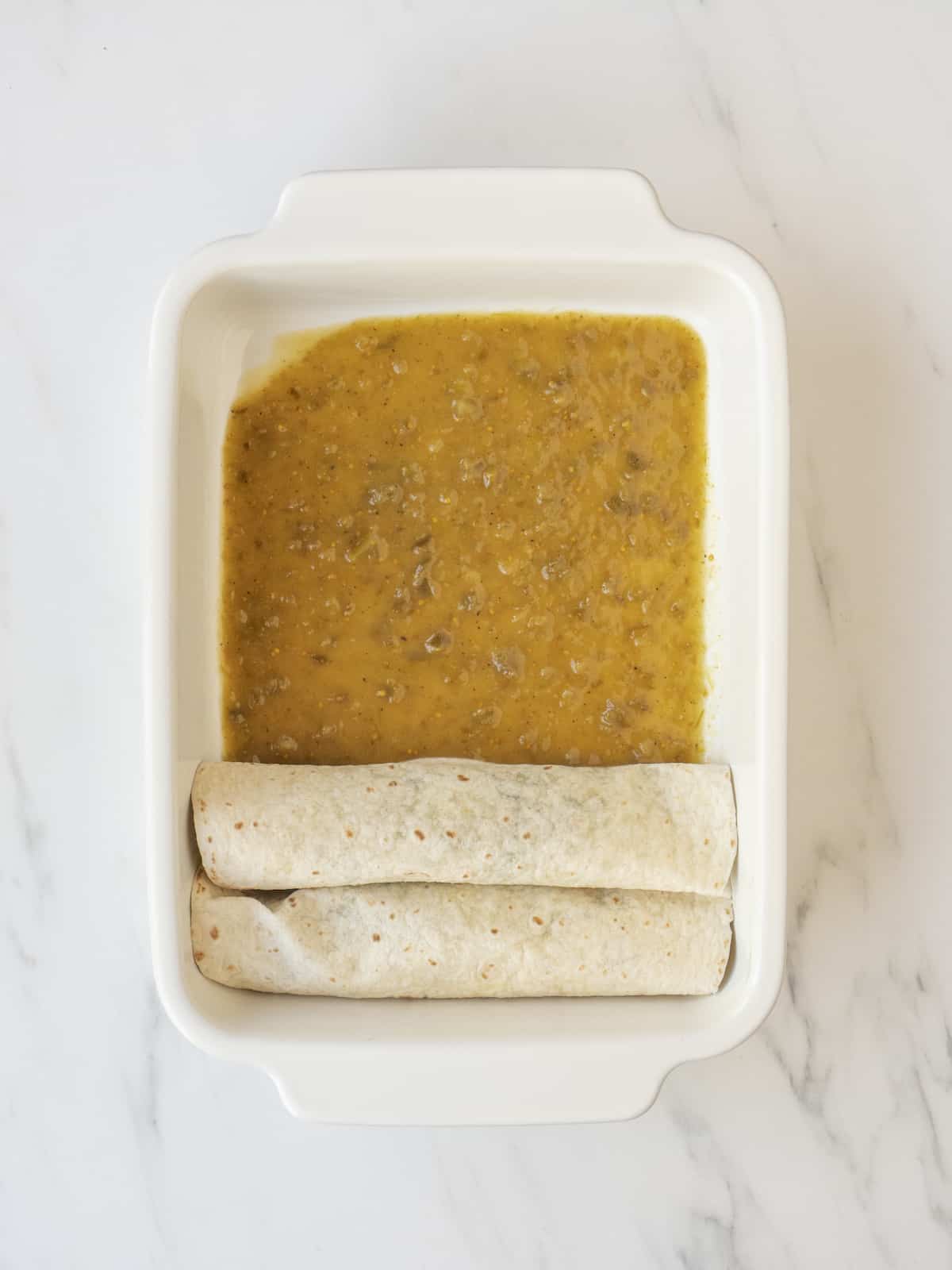 A white rectangular baking dish with enchilada sauce in the bottom of the dish and two vegetarian tortillas filled and rolled up placed with the seam down.