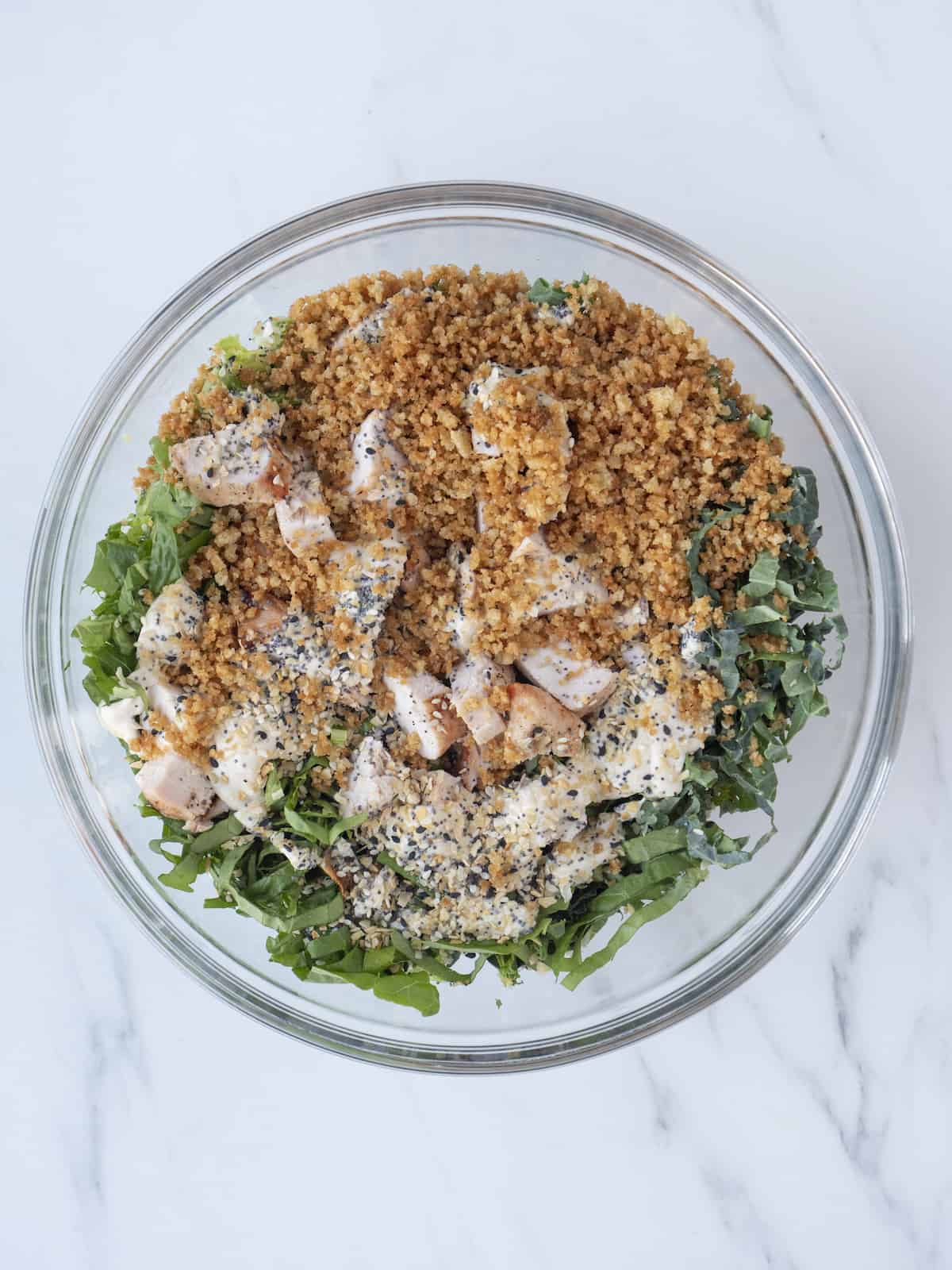 A large glass mixing bowl with ingredients for chicken caesar wrap filling, shredded greens, grilled chicken, bread crumbs and tahini dressing.