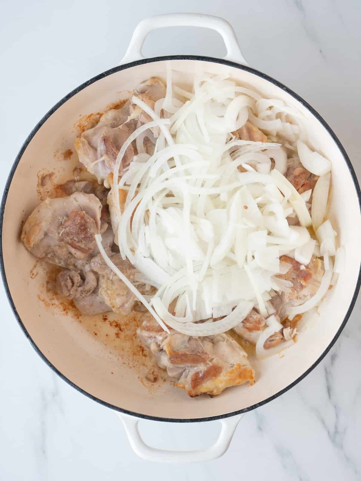A white dutch oven with seared chicken thighs and thinly sliced onions just added.