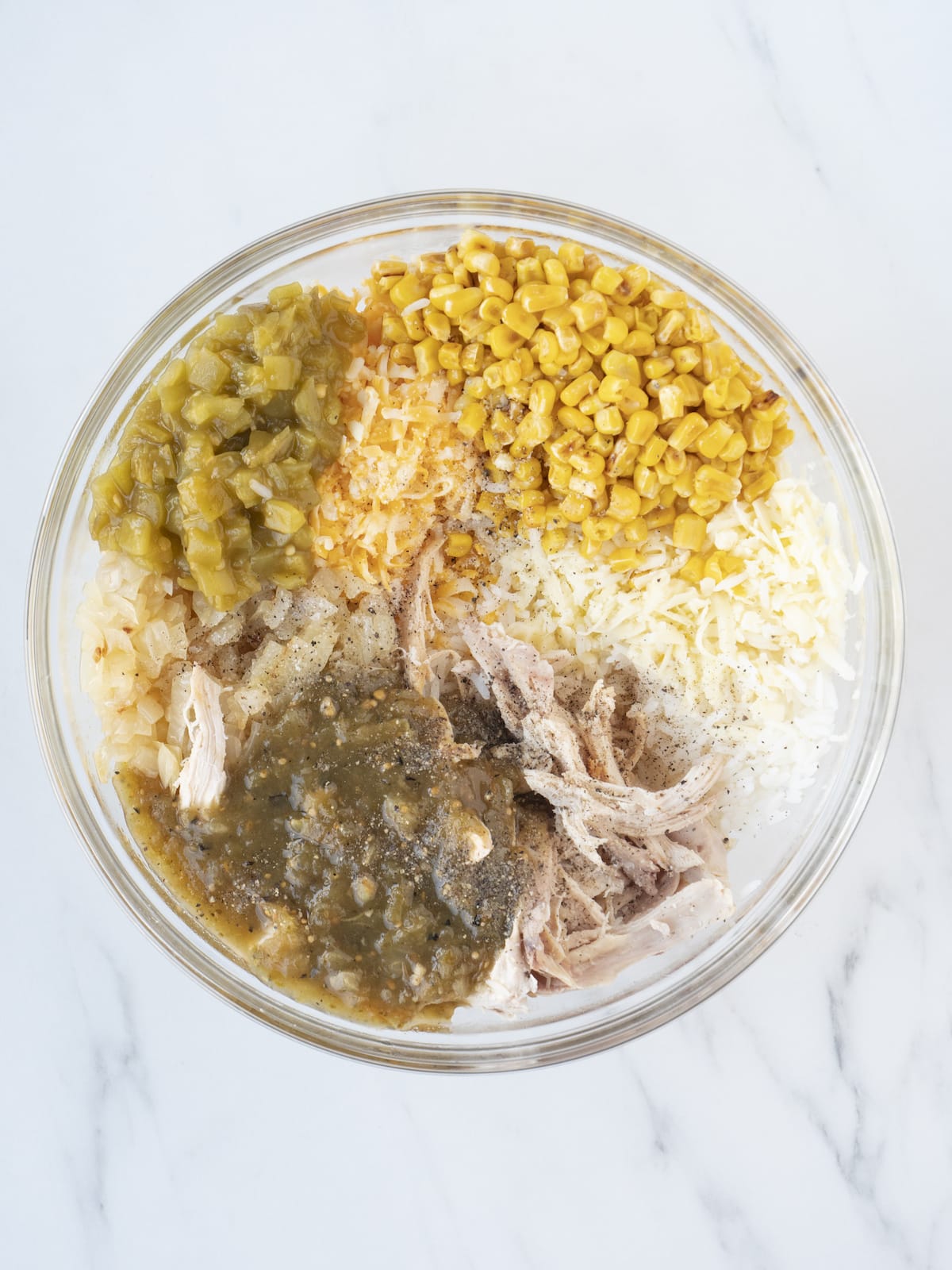 A large glass mixing bowl, with cooked rice in the bottom, topped with corn, chopped onions, shredded mozzarella and colby jack cheeses, shredded chicken, green chiles and tomatillo salsa, sprinkled with salt and black pepper.