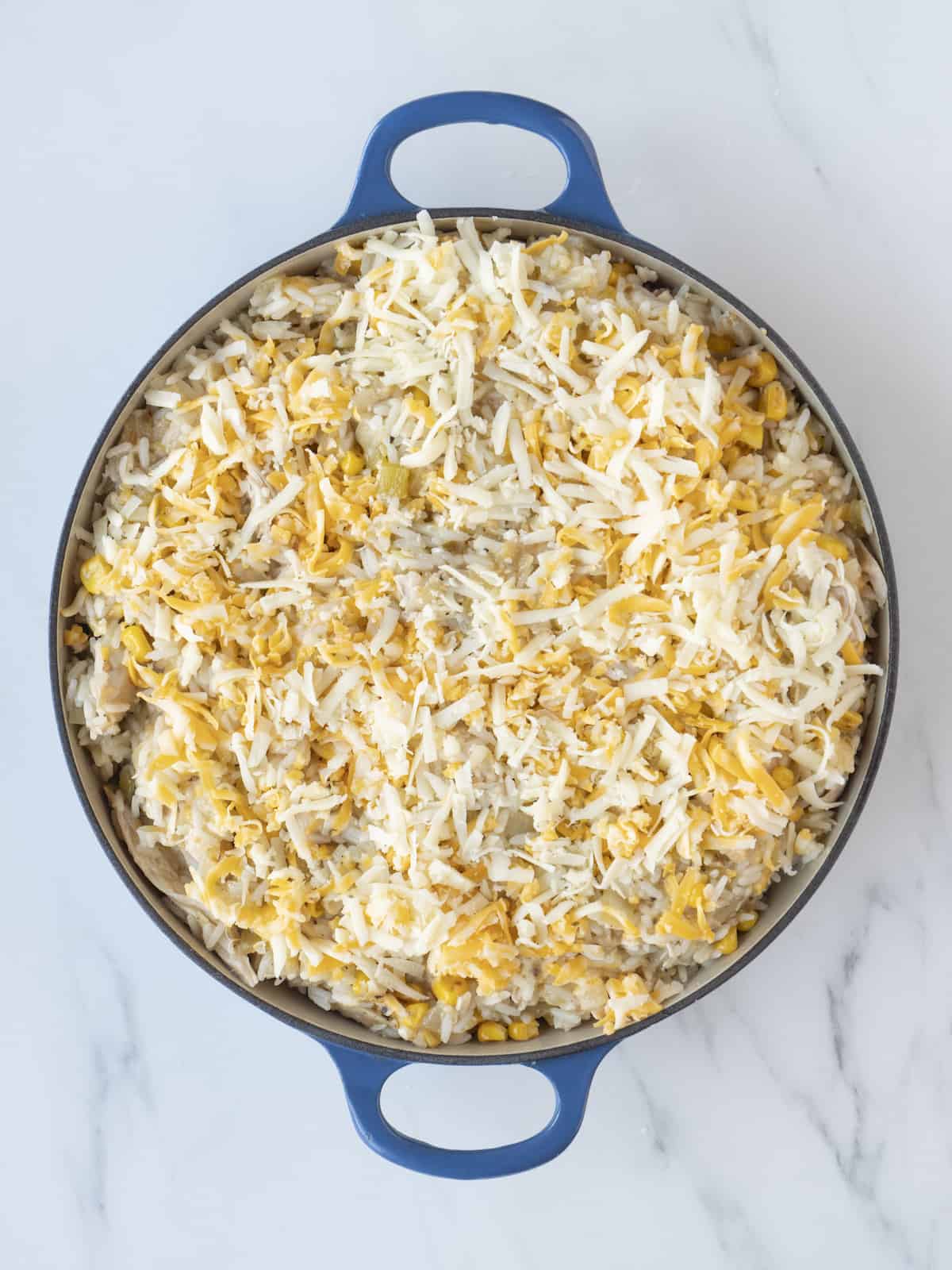 A blue dutch oven with a chicken and rice casserole assembled, with refried beans in the bottom, topped with the mixture of chicken, rice, veggies and cheese, topped with shredded cheese.