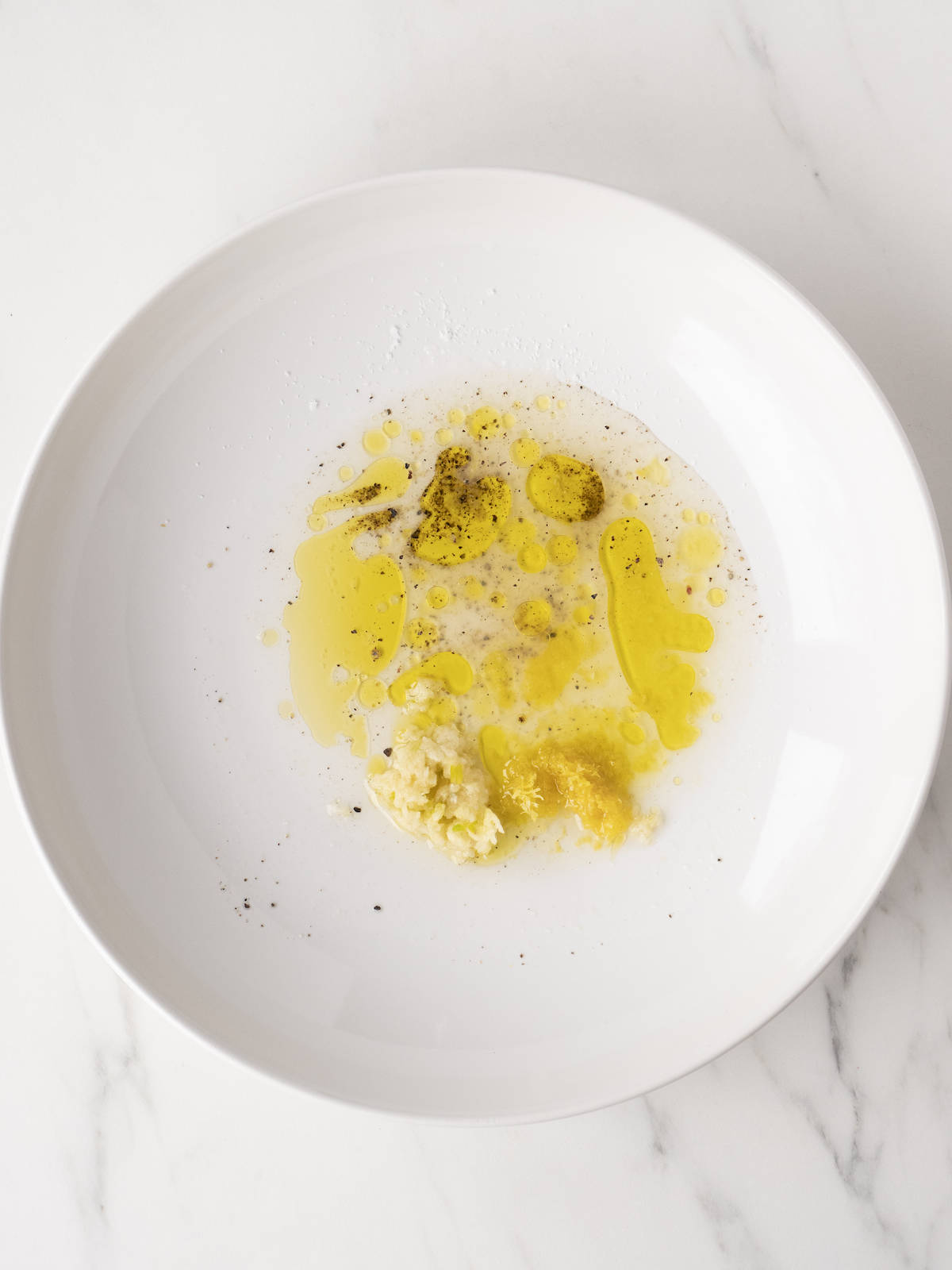 A large white bowl with ingredients for dressing in it, olive oil, garlic, lemon zest and juice, salt and pepper.