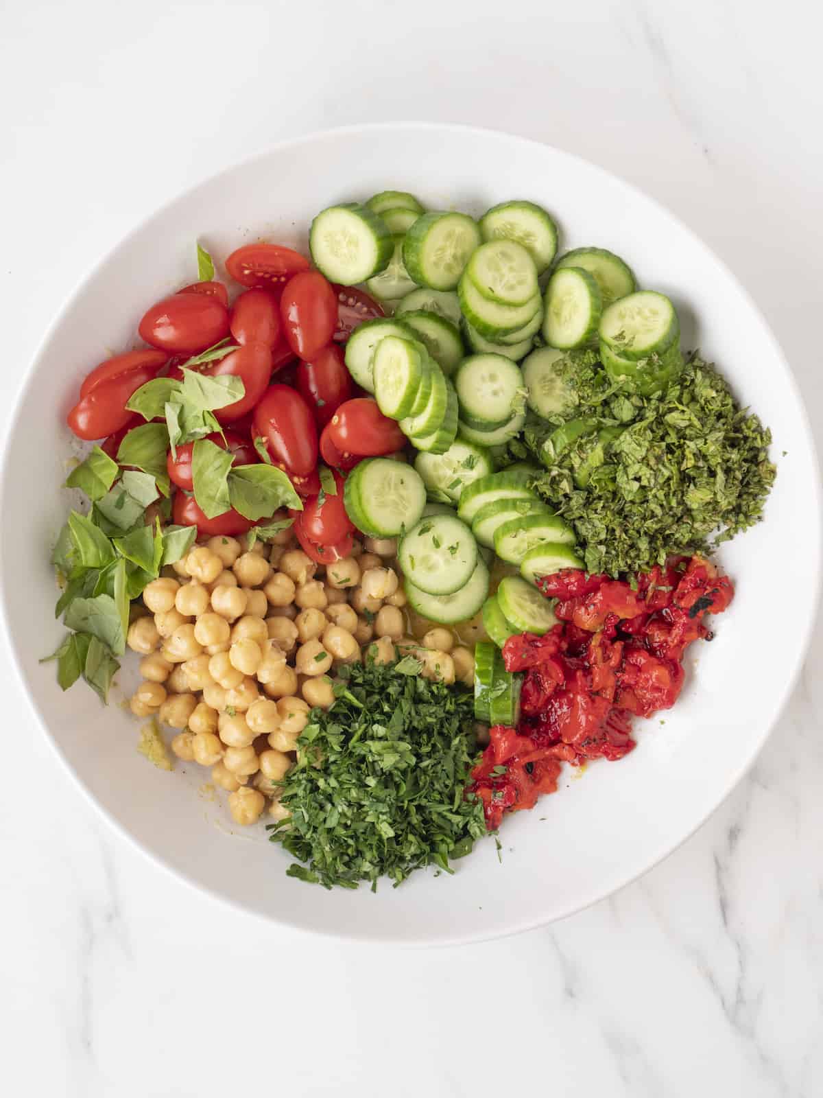 A large white bowl with dressing at the bottom, and salad ingredients on top, tomatoes, slices cucumbers, chopped roasted red pepper, chickpeas, chopped mint and parsley and basil.