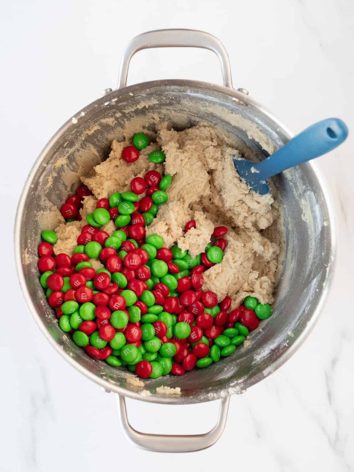 A stand mixer bowl with cookie dough, red and green M&Ms just added and a blue spatula. 