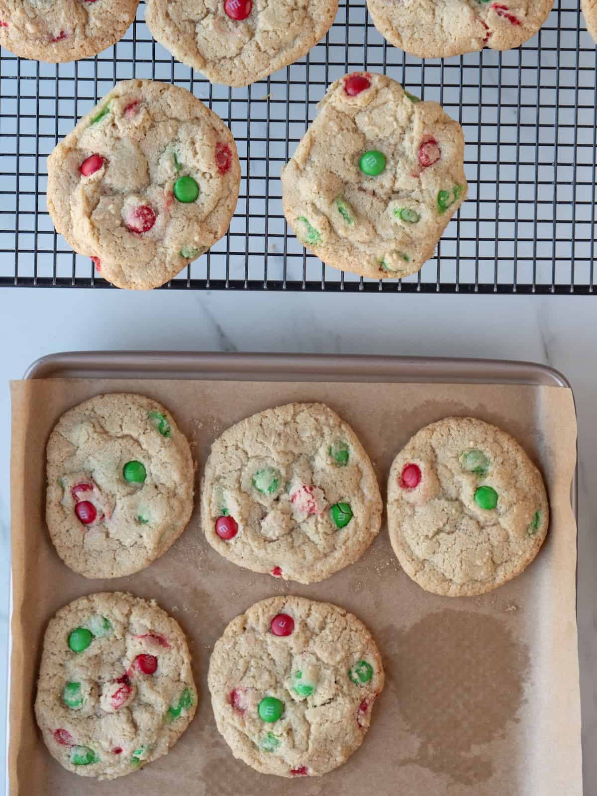 A wire rack alongside a parchment lined baking sheet, both with Christmas M&M cookies.