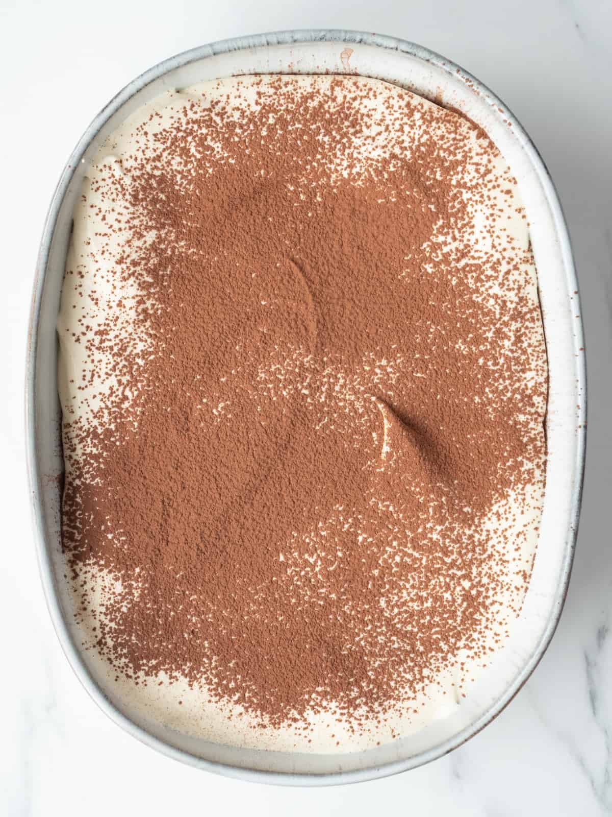 An oval platter with tiramisu being constructed in it, with one layer of ladyfingers topped with mascarpone-cream mixture dusted with cocoa powder.