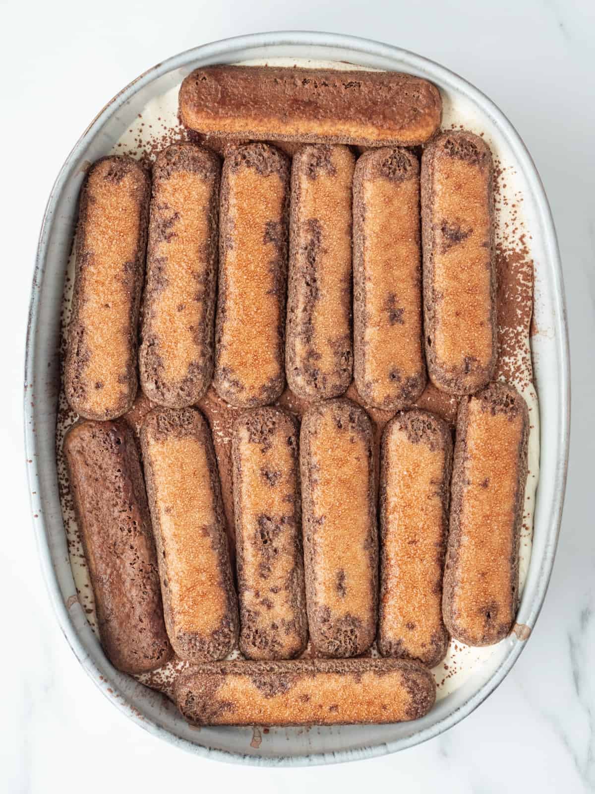 An oval platter with a second layer of espresso mixture soaked ladyfingers on top of an already layered ladyfinger and mascarpone mixture layer in order to construct a tiramisu.