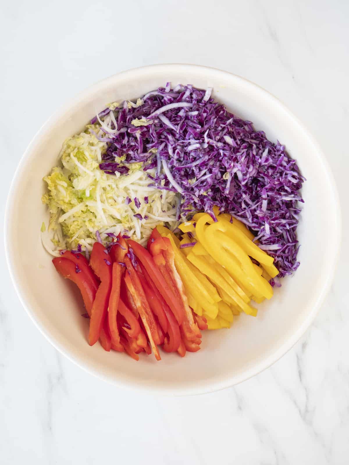 A white bowl with shredded napa and purple cabbages, and red and yellow peppers cut into matchsticks.