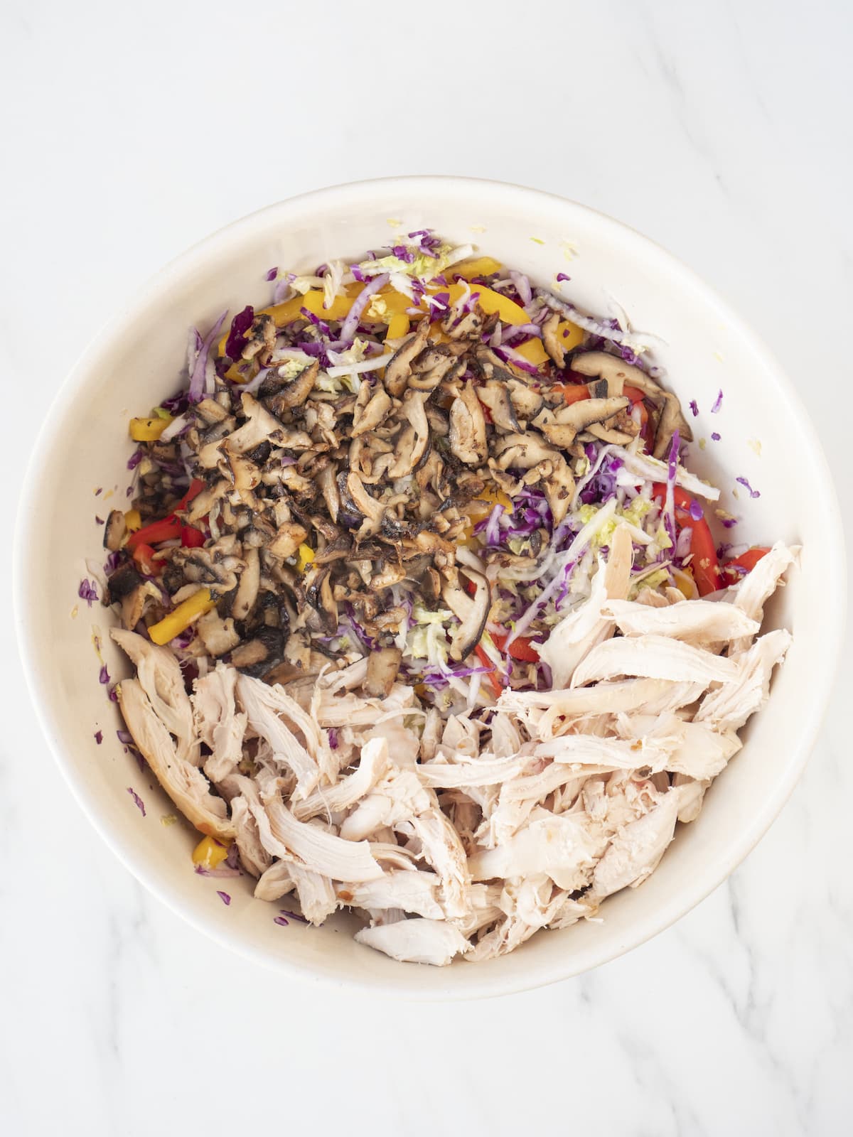A white bowl with all the veggies- cabbages, bell peppers and mushrooms mixed in, along with shredded chicken breasts.