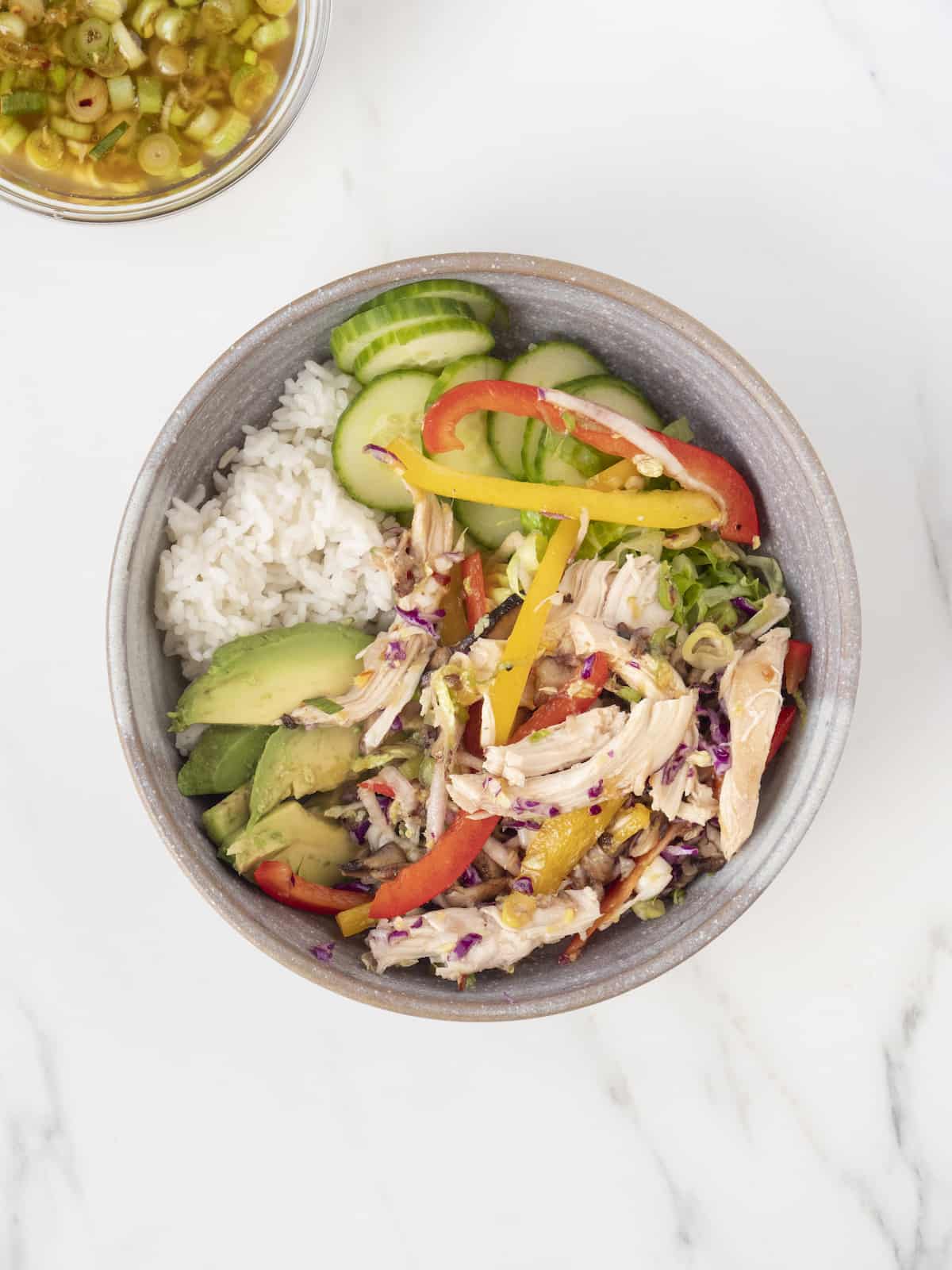 A grey ceramic bowl with white rice at the bottom, topped with the chicken-vegetable mixture, sliced avocados, shredded lettuce and sliced cucumbers.
