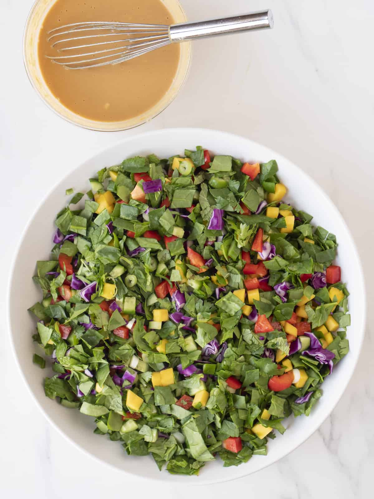 A large white bowl of all the chopped vegetables, herbs and mango tossed  together for the mango cucumber chopped salad, with a small glass bowl of peanut-soy dressing with a whisk on the side.