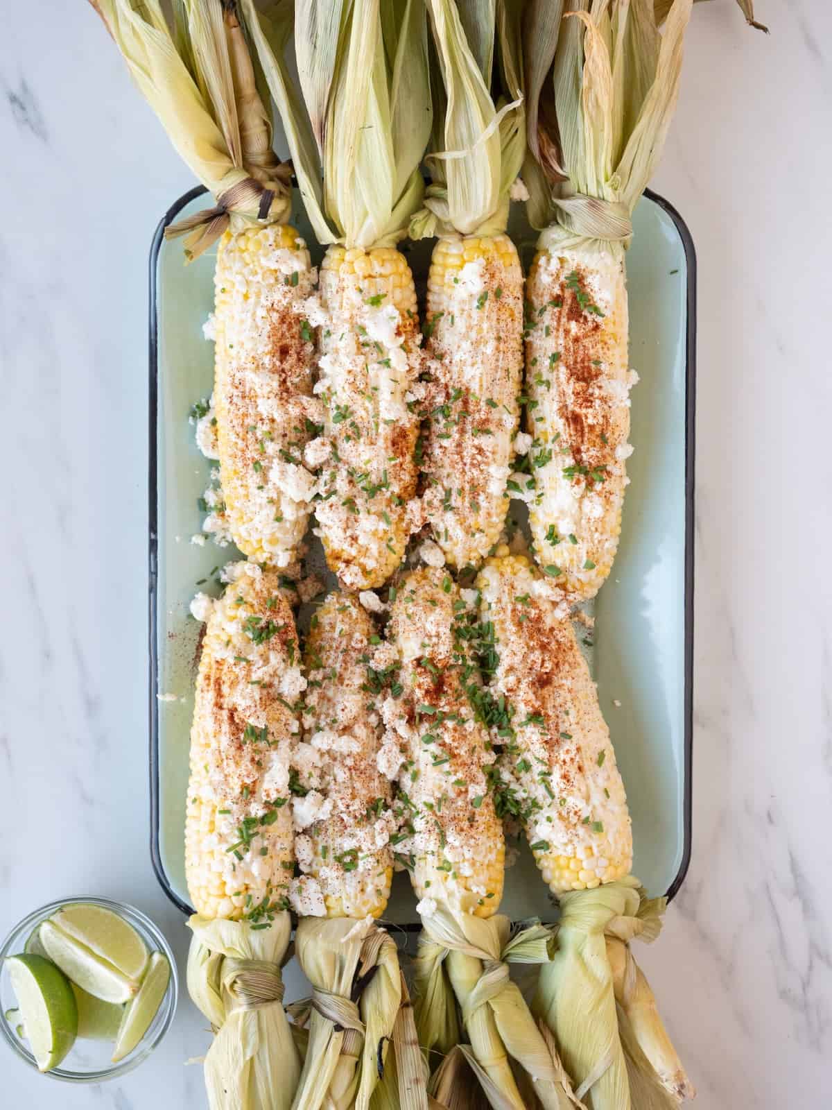 A blue and green rectangular platter with 8 corn on the cob placed with the husks pulled back but still attached, brushed with mayo and topped with chili pepper mixture, crumbled feta and chives, along with a small bowl of lime wedges on the side.