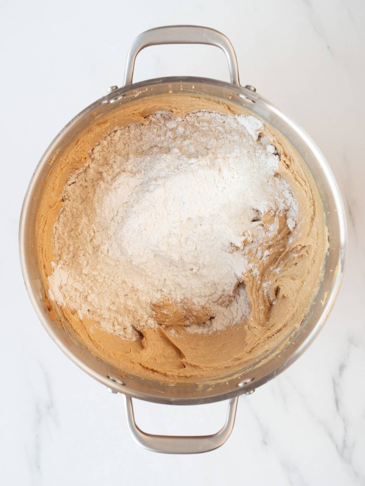 A stand mixer bowl with a smooth mixture of all wet ingredients and sugars, with a mix of flour and baking soda added to it.