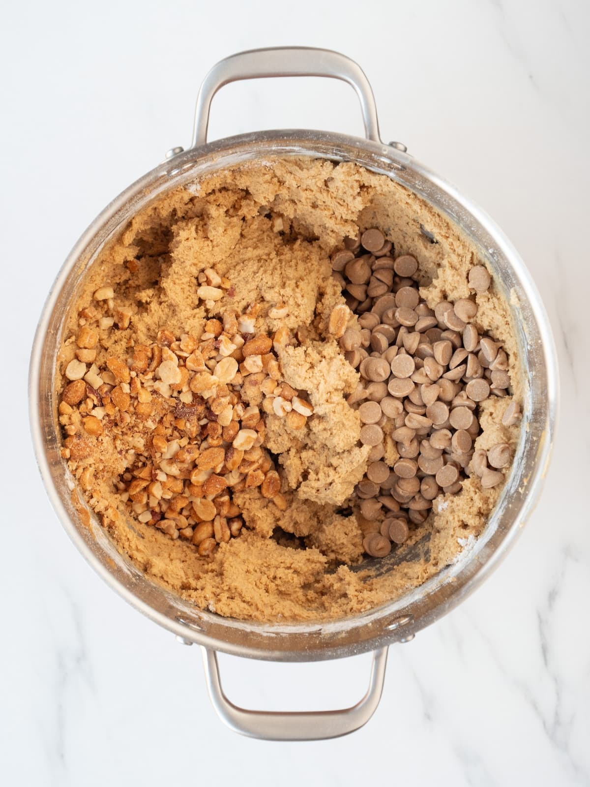 A stand mixer bowl with peanut butter cookie dough and peanut butter chips and peanuts added to it. 