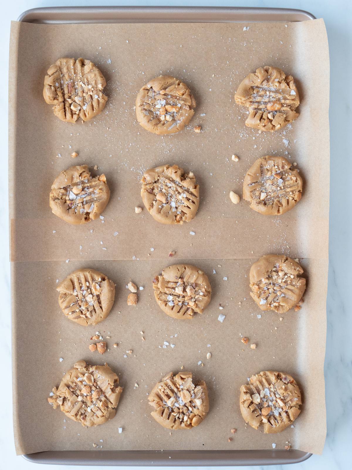 A parchment-lined baking sheet with scoops of peanut butter cookie dough flattened with a fork leaving a crisscross pattern on them, topped with Maldon salt and ready to be baked.