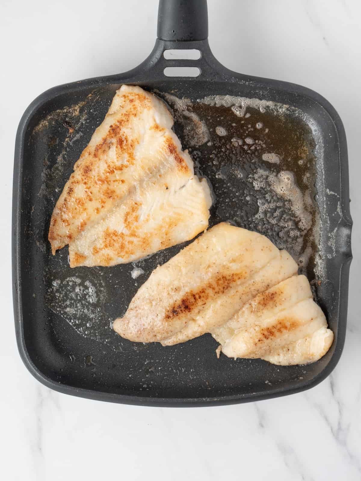 A nonstick skillet with butter, and two sole fillets cooked on the top side already and being cooked on the other side.