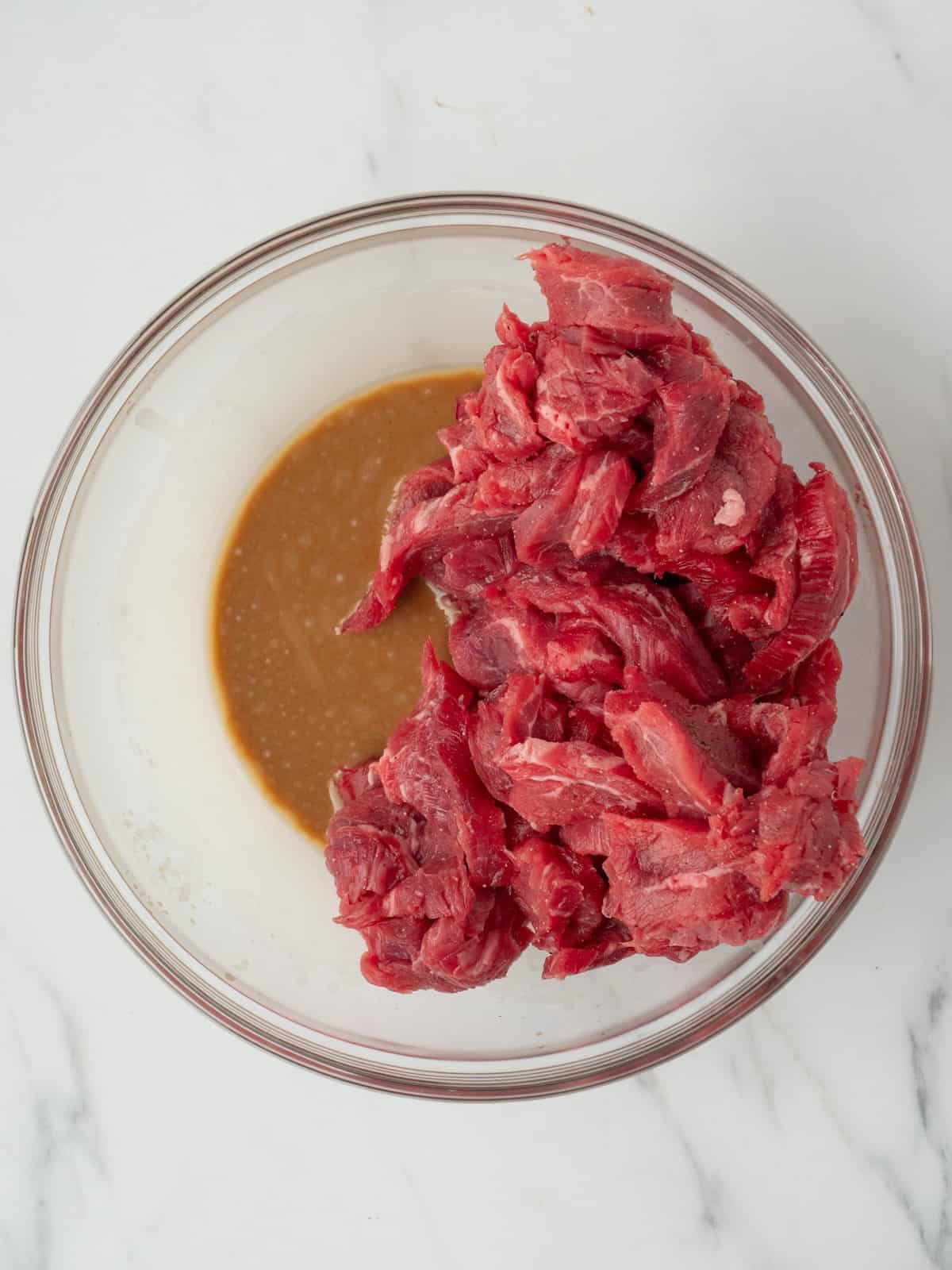 A glass mixing bowl with the steak strips and the soy sauce mixture, in order to coat the steak strips with the mixture.