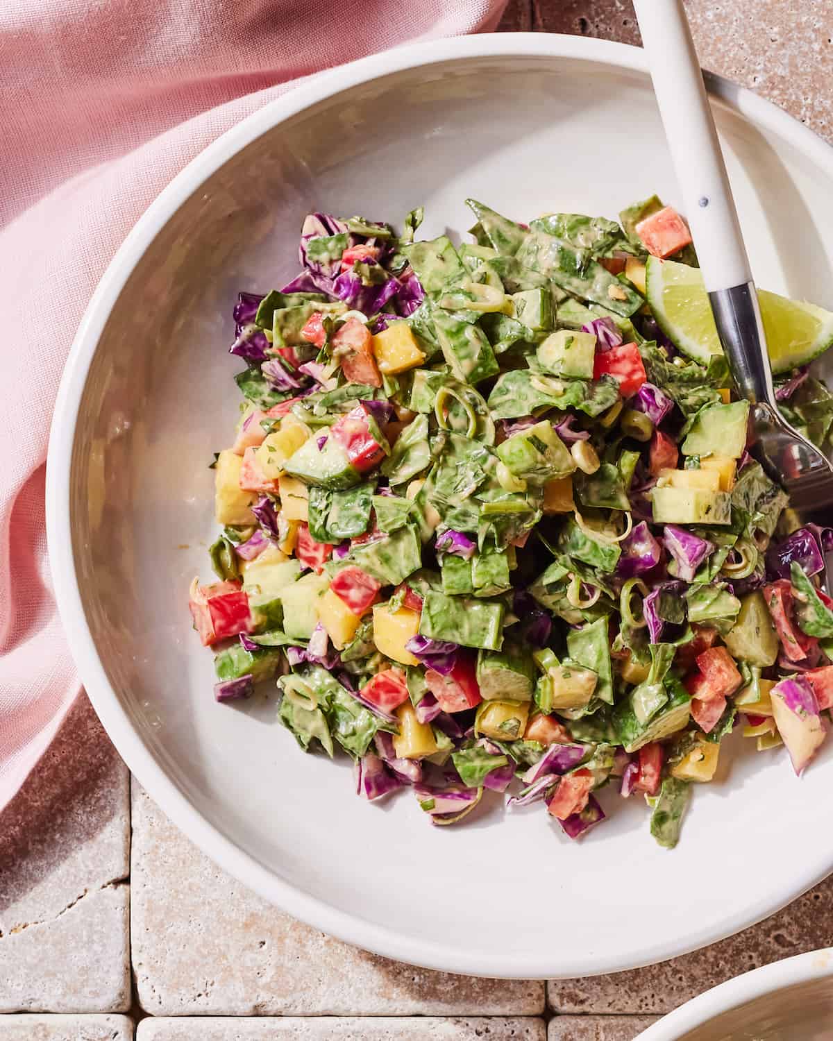 A white low bowl of mango cucumber chopped salad and a lime wedge, with a fork in it, placed on a beige tiled surface with a pink cloth napkin on the side.