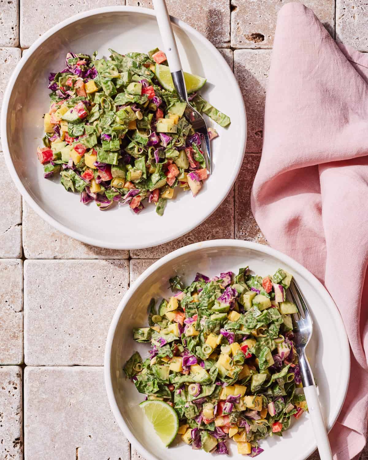 Two white low bowls of mango cucumber chopped salad, with a fork and a lime wedge in each bowl, placed on a beige tiled surface with a pink cloth napkin on the side.