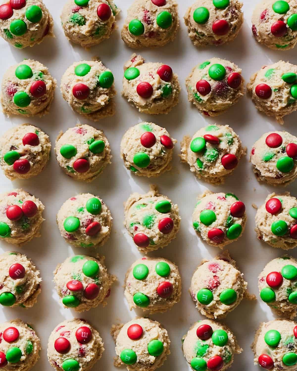 A parchment-lined baking sheet with scoops of Christmas M&M cookie dough, with more red and green M&Ms stuck on top of the scoop.