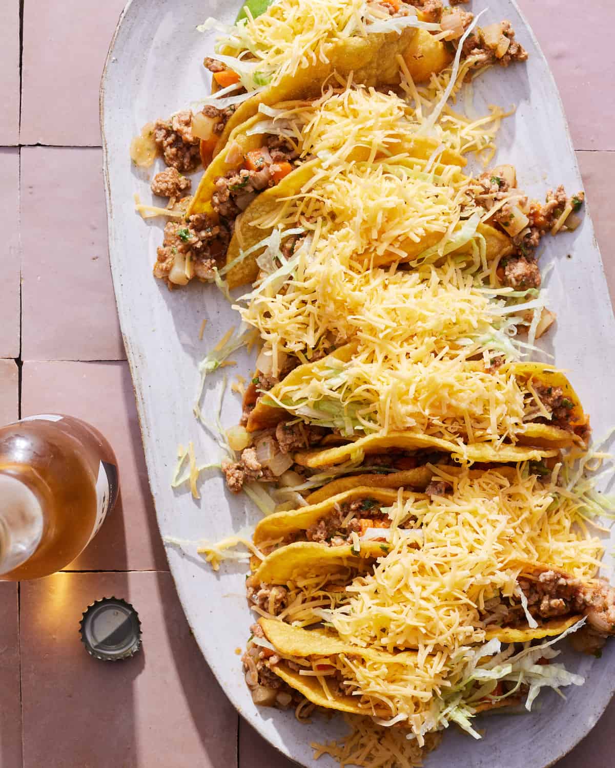 An oval white platter with picadillo hard shell tacos topped with shredded cheese, placed on a tiled surface, with an open beverage in a bottle on the side.
