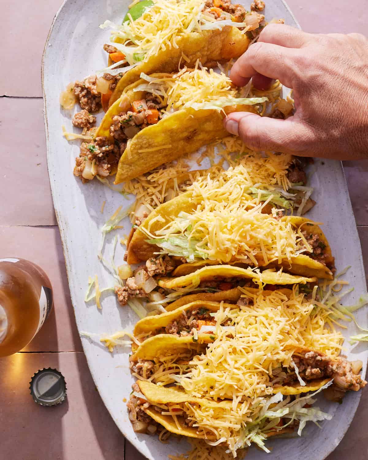 An oval white platter with picadillo hard shell tacos topped with shredded cheese, placed on a tiled surface, with an open beverage in a bottle on the side, with a hand picking up a taco from the platter.