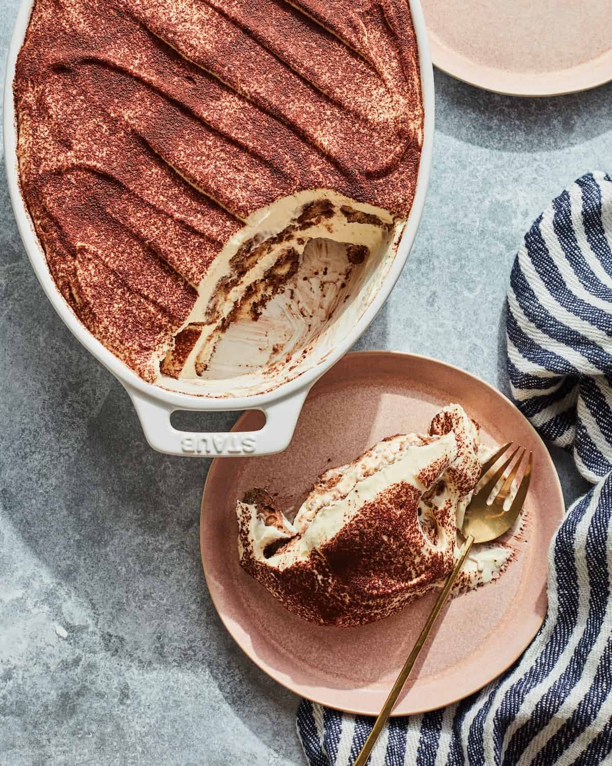 A white oval dish with tiramisu, one corner of which has been served into a small ceramic pink plate on the side, with a golden fork in the plate, with a navy and white striped kitchen towel on the side.