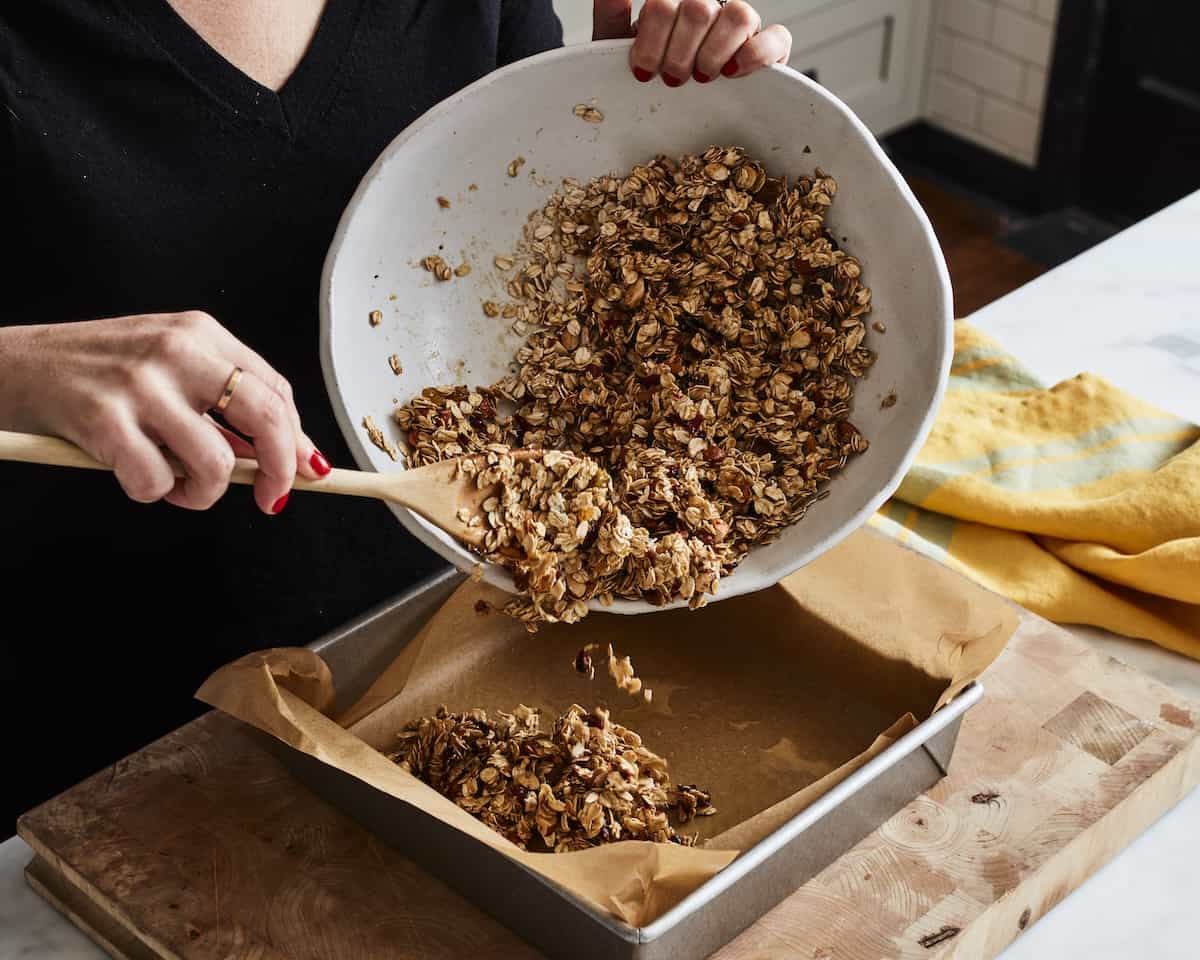 A woman pouring the homemade granola bar mixture from a white ceramic bowl into a 9x9 square parchment paper lined baking pan using a wooden spoon.