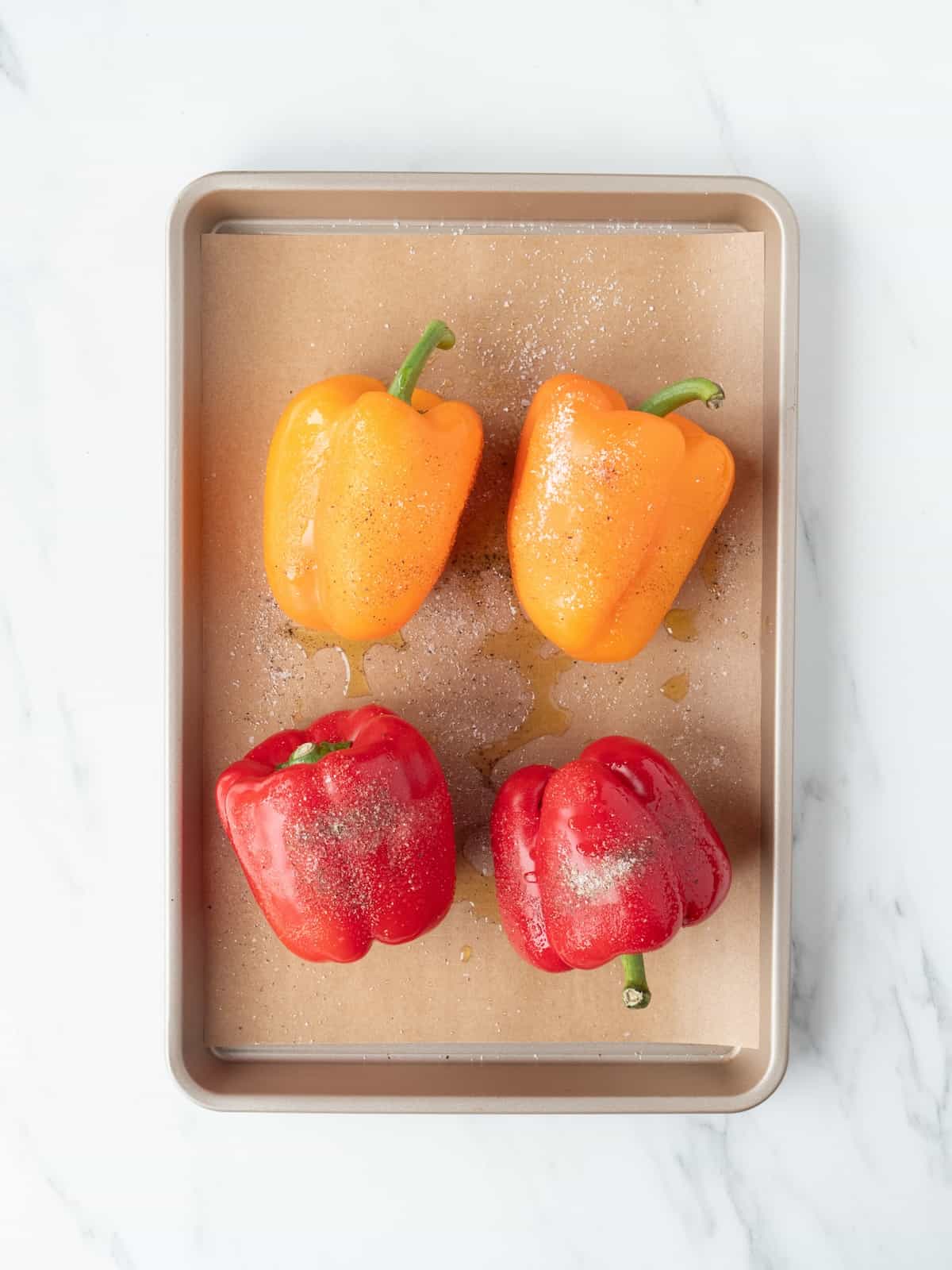 A rectangular baking sheet lined with parchment paper with two red and two yellow bell peppers tossed in oil and sprinkled with salt and pepper.