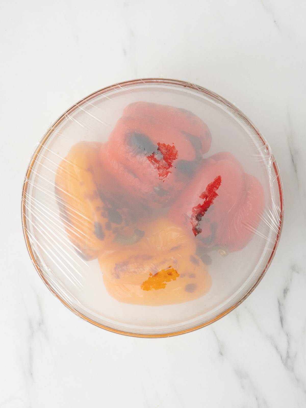 A glass mixing bowl with roasted red and yellow bell peppers, and the bowl is covered with cling wrap.