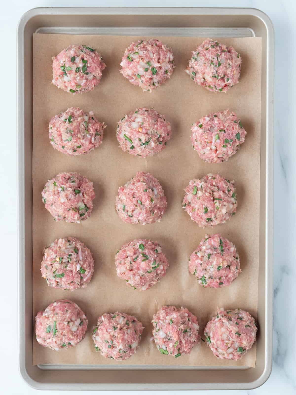 A parchment-lined baking sheet with pork meatballs placed in a 3 by 5 grid, shaped in spheres, ready to be baked.