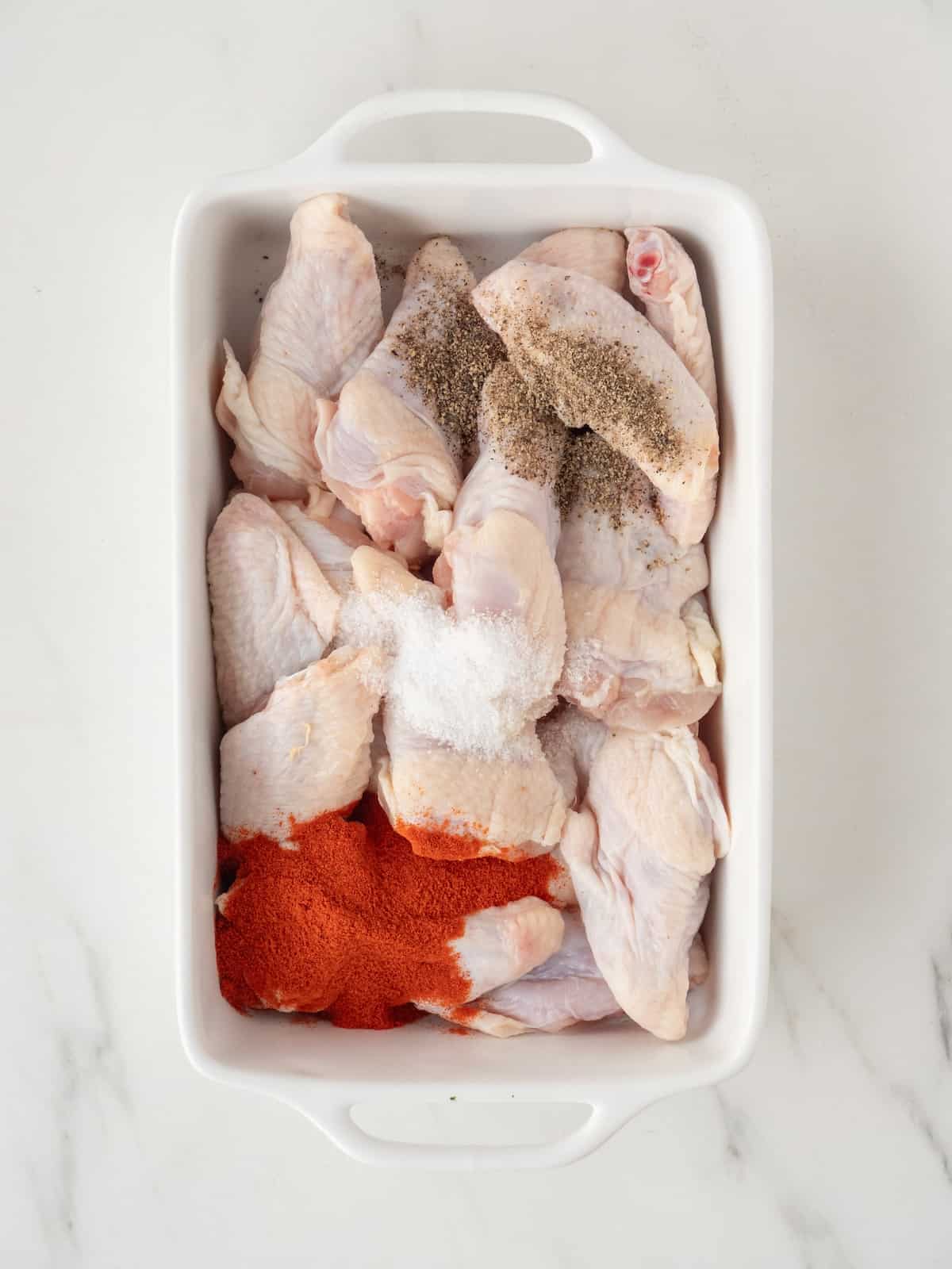 A white rectangular ceramic dish with raw chicken wings topped with paprika, salt and pepper.
