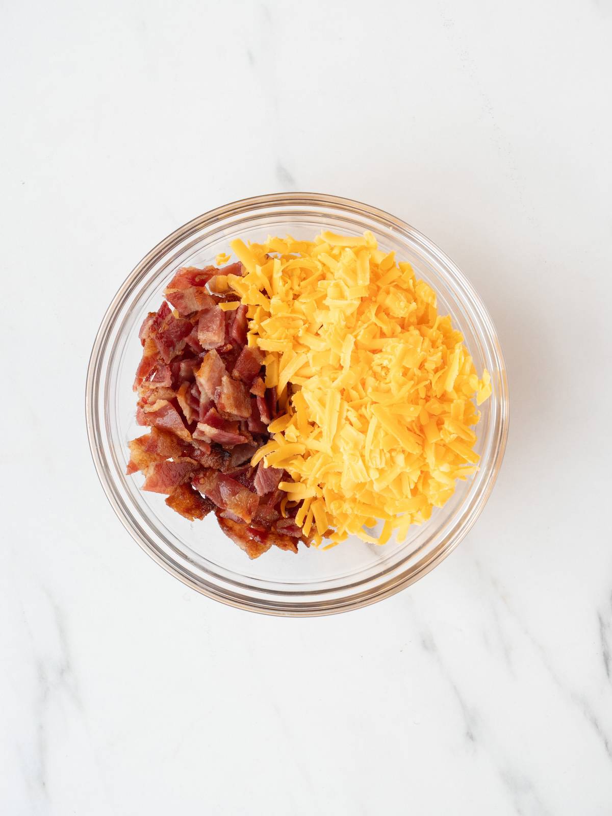 A glass mixing bowl with bacon broken into pieces and shredded cheddar cheese.
