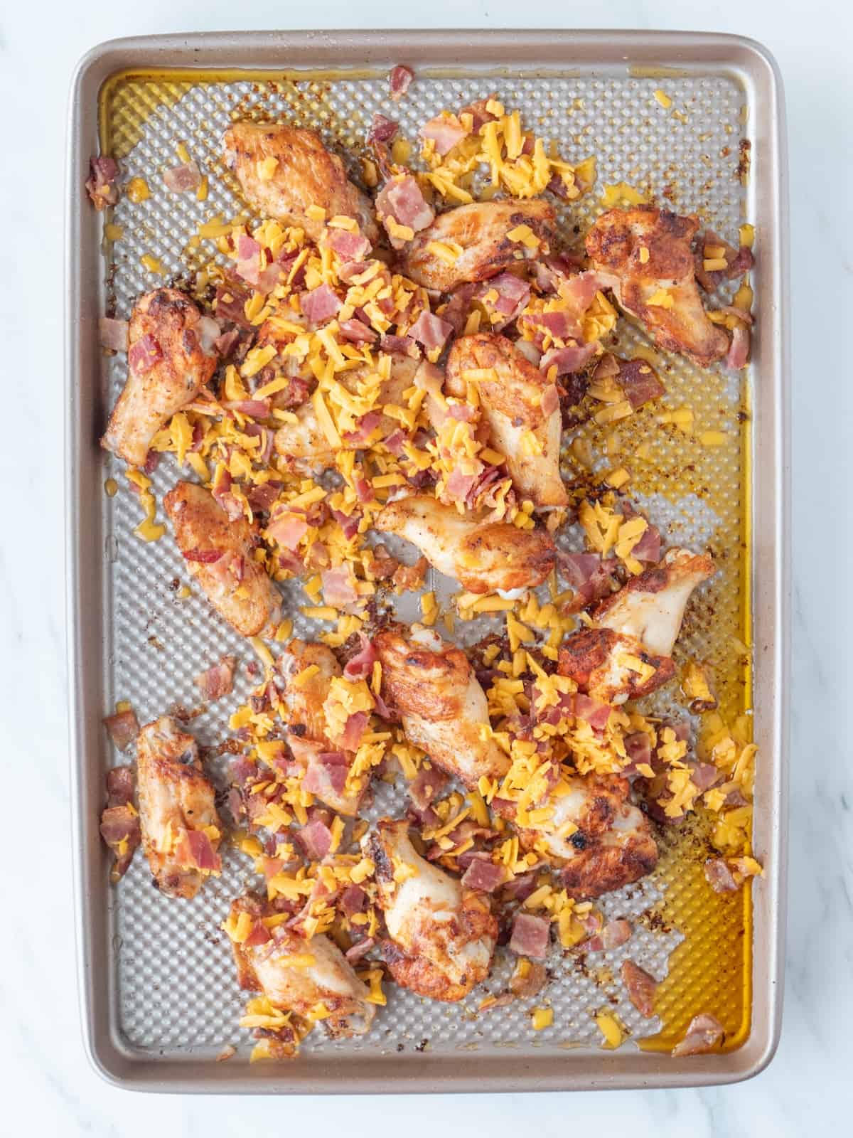 A baking sheet with chicken wings baked in the oven, topped with bacon pieces and shredded cheese.