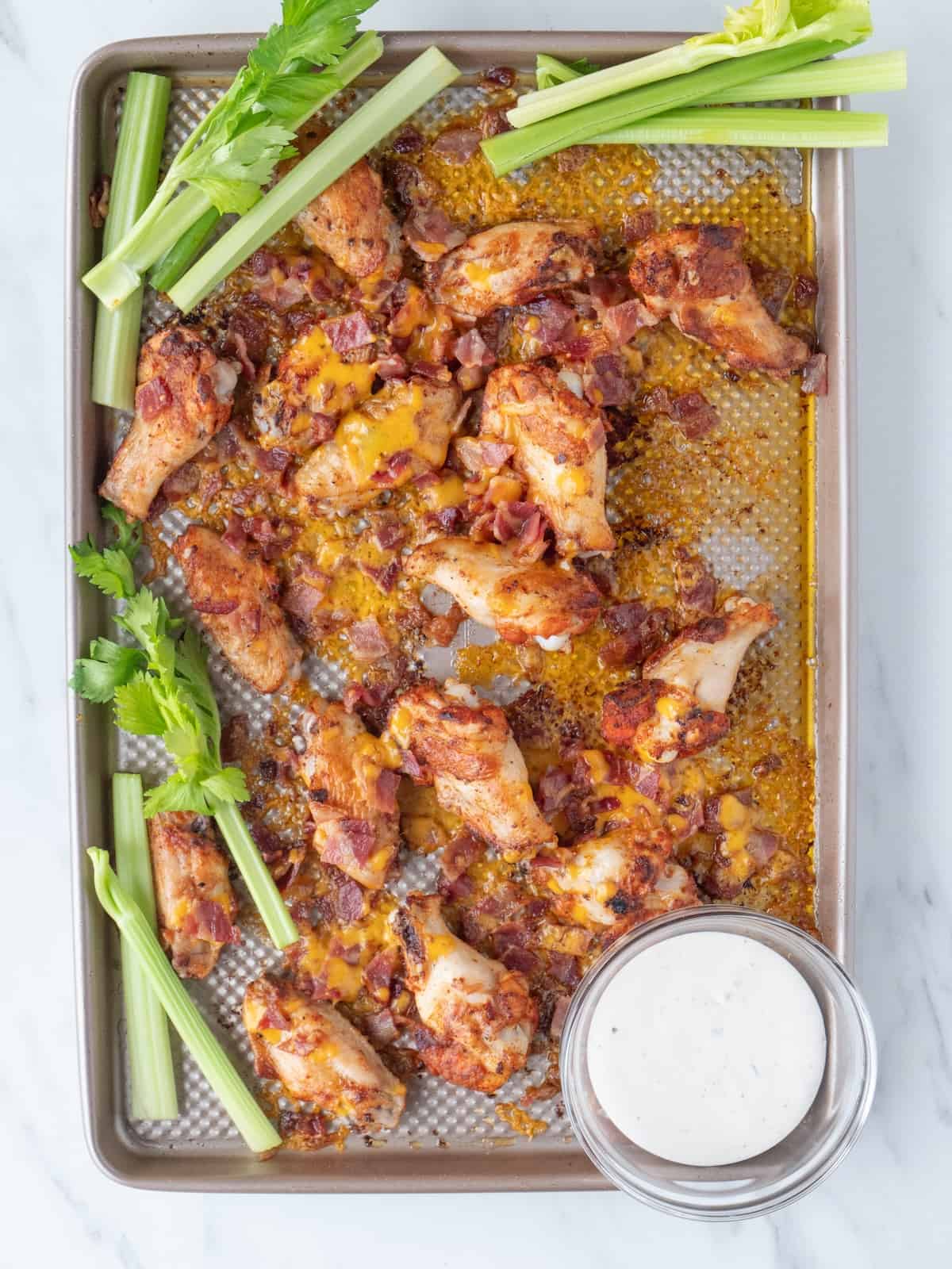 A baking sheet with chicken wings topped with bacon pieces and melted cheese, fresh out of the oven, along with celery sticks and a little glass bowl of ranch in the corner.
