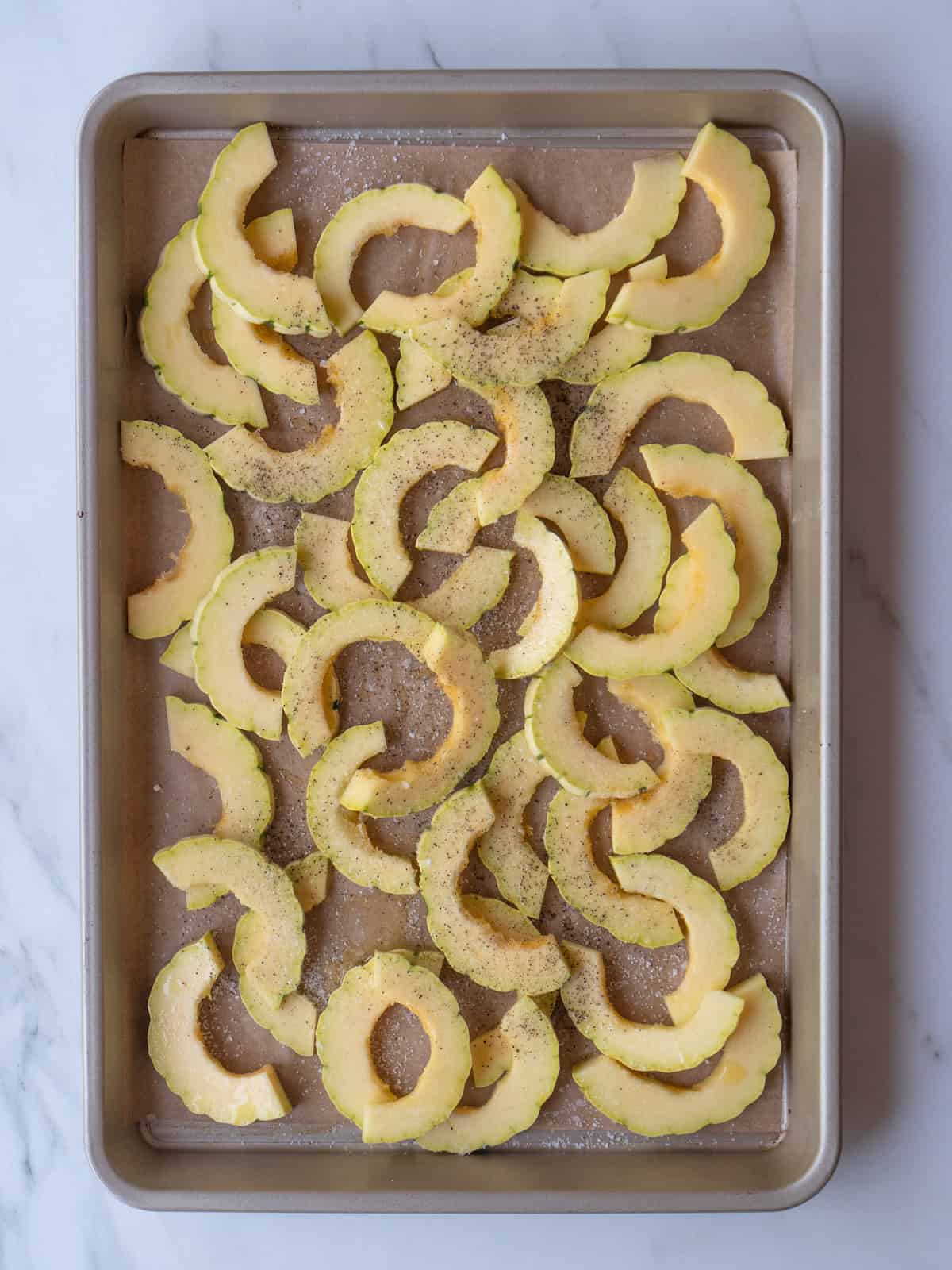 A baking sheet lined with parchment paper with sliced delicata squash sprinkled with salt and pepper, drizzled with olive oil. 