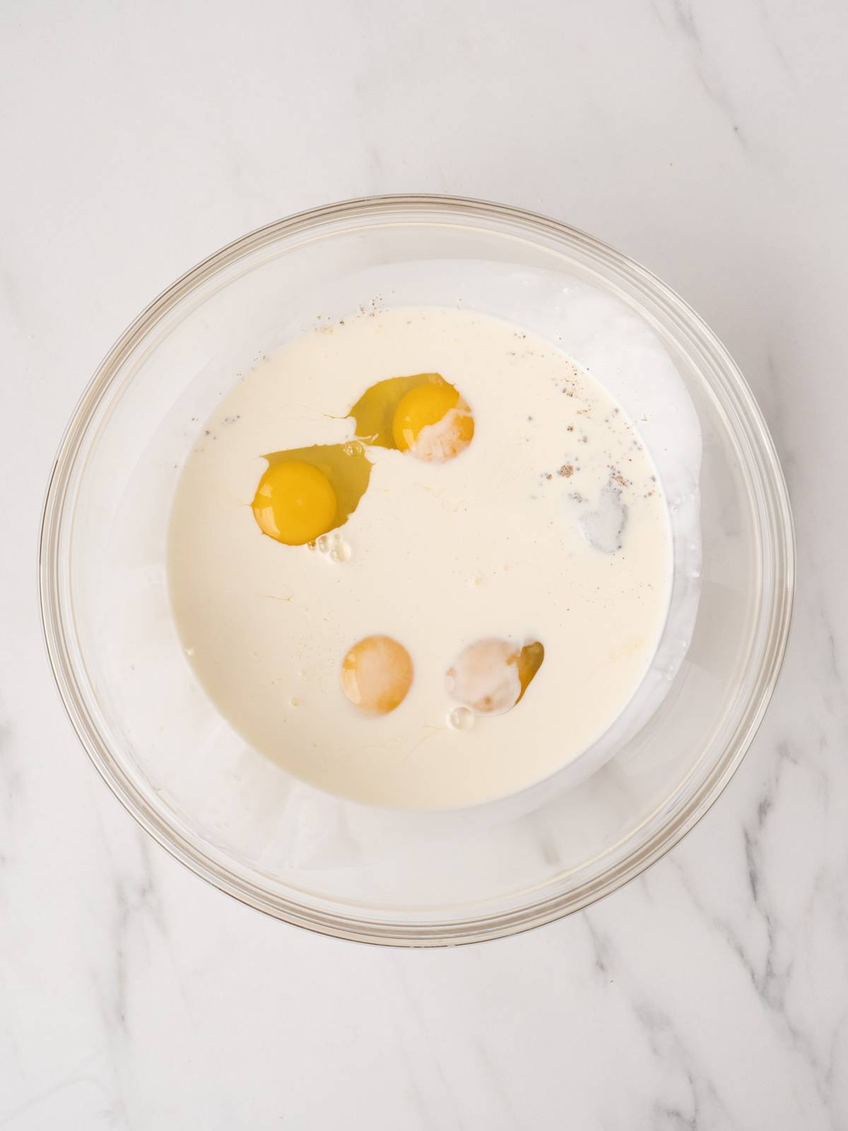 A large glass mixing bowl with cream, milk, eggs, salt and pepper.
