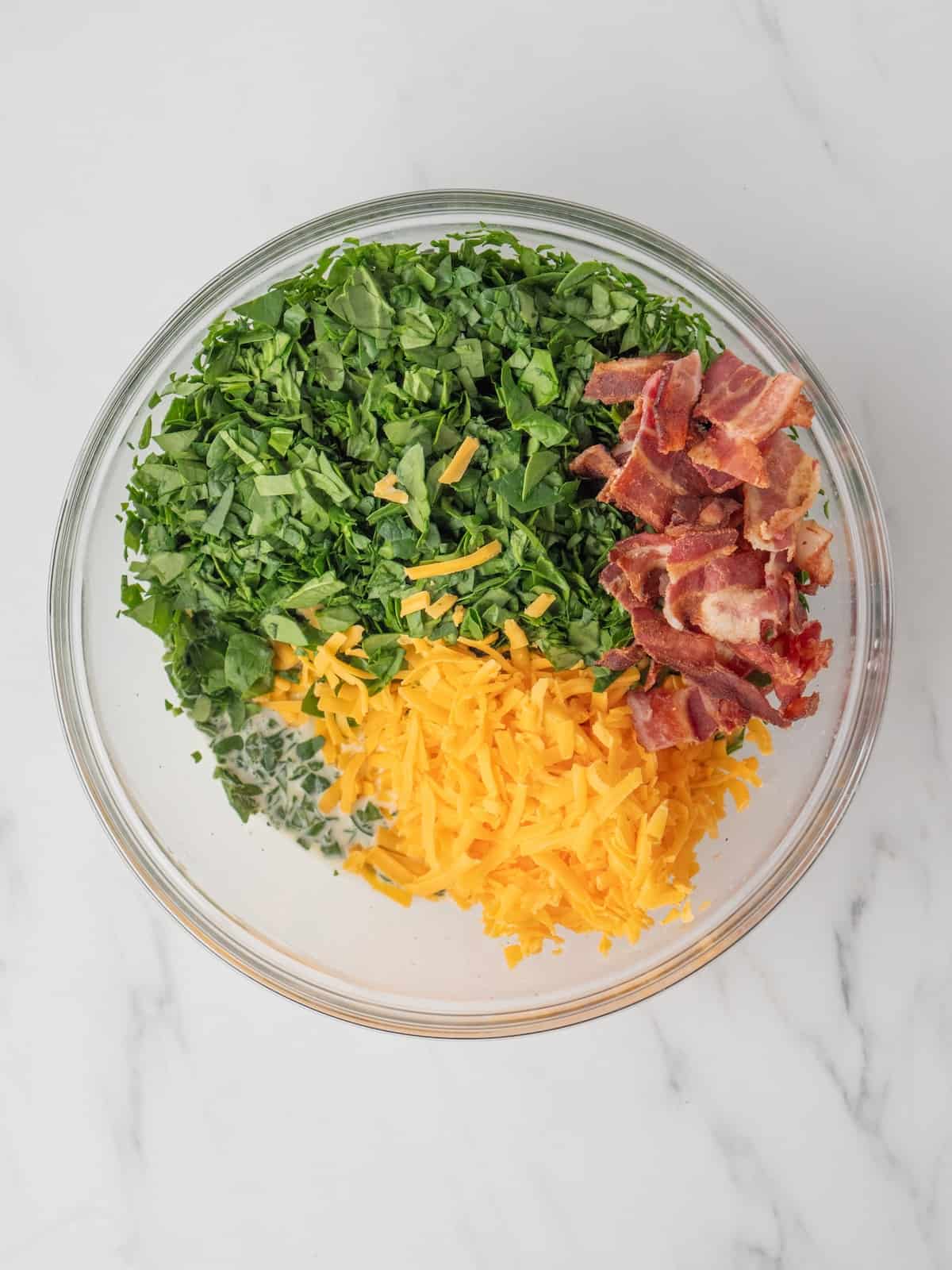 A large glass mixing bowl with an egg, milk and cream mixture topped with torn bacon, shredded cheese and chopped spinach.